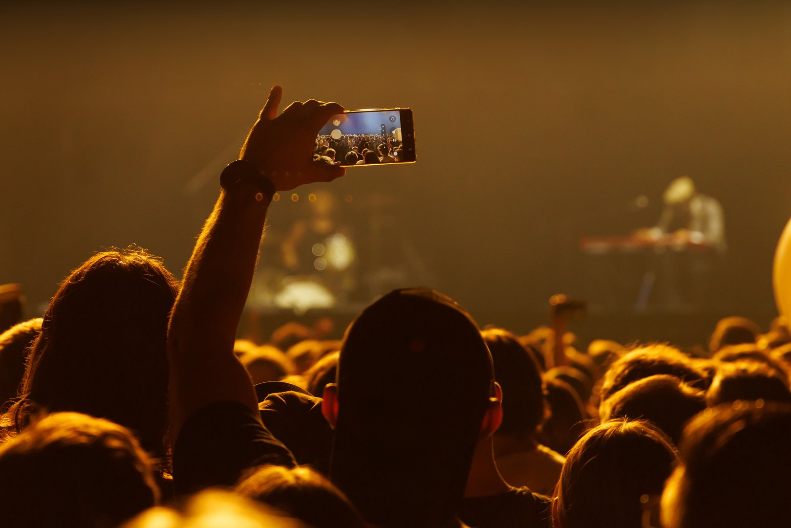 A person captures a live music concert using a smartphone with orange-golden lighting around the crowd. The image expresses energy, modernity, and collective enjoyment of live performances. Ideal for lifestyle, music, and technology uses.