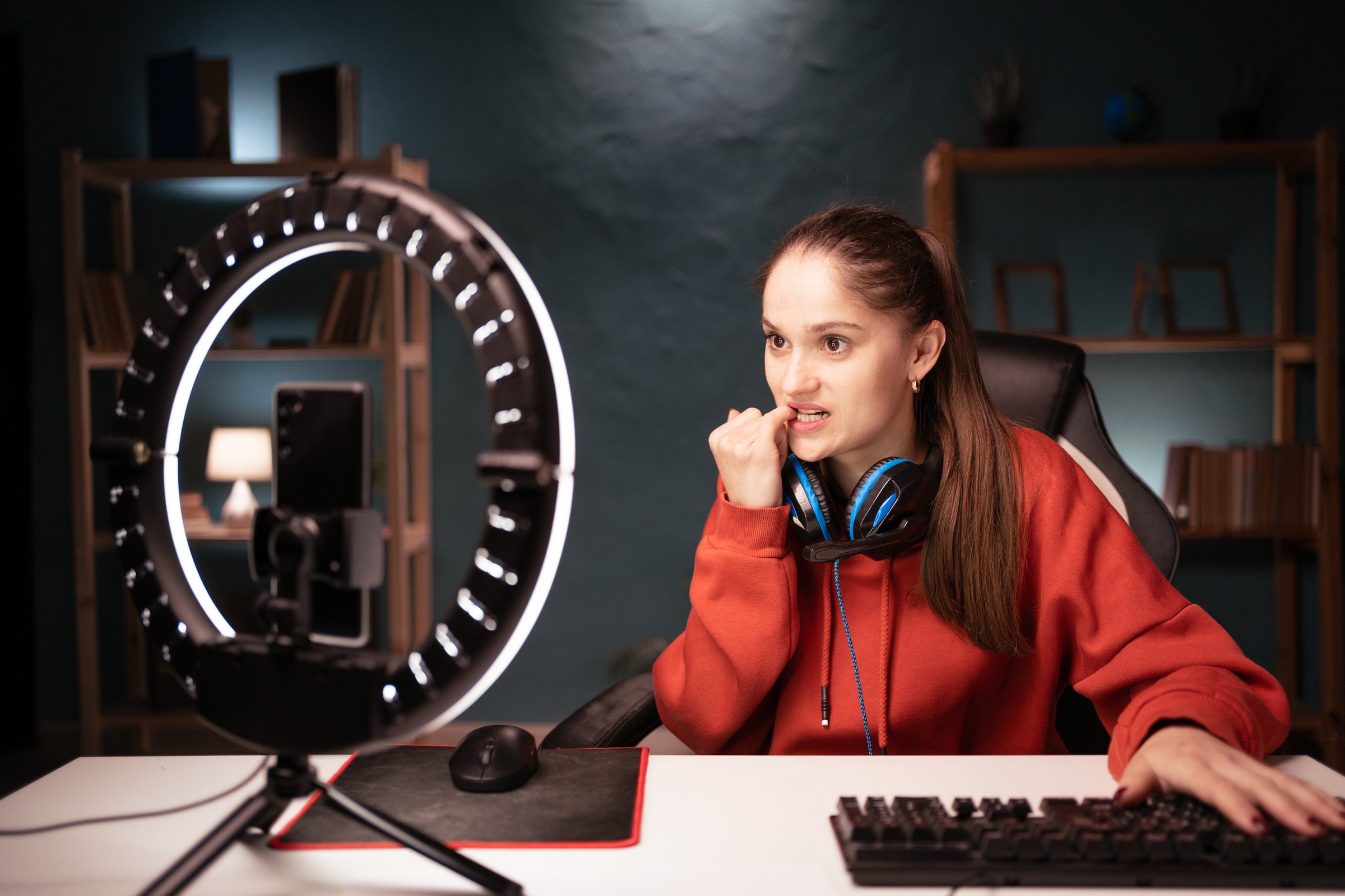 Woman gamer blogger influencer streaming live with her subscribers, sharing online her cybersport game using smartphone and ring lamp, nervous worried biting nails. Copy space