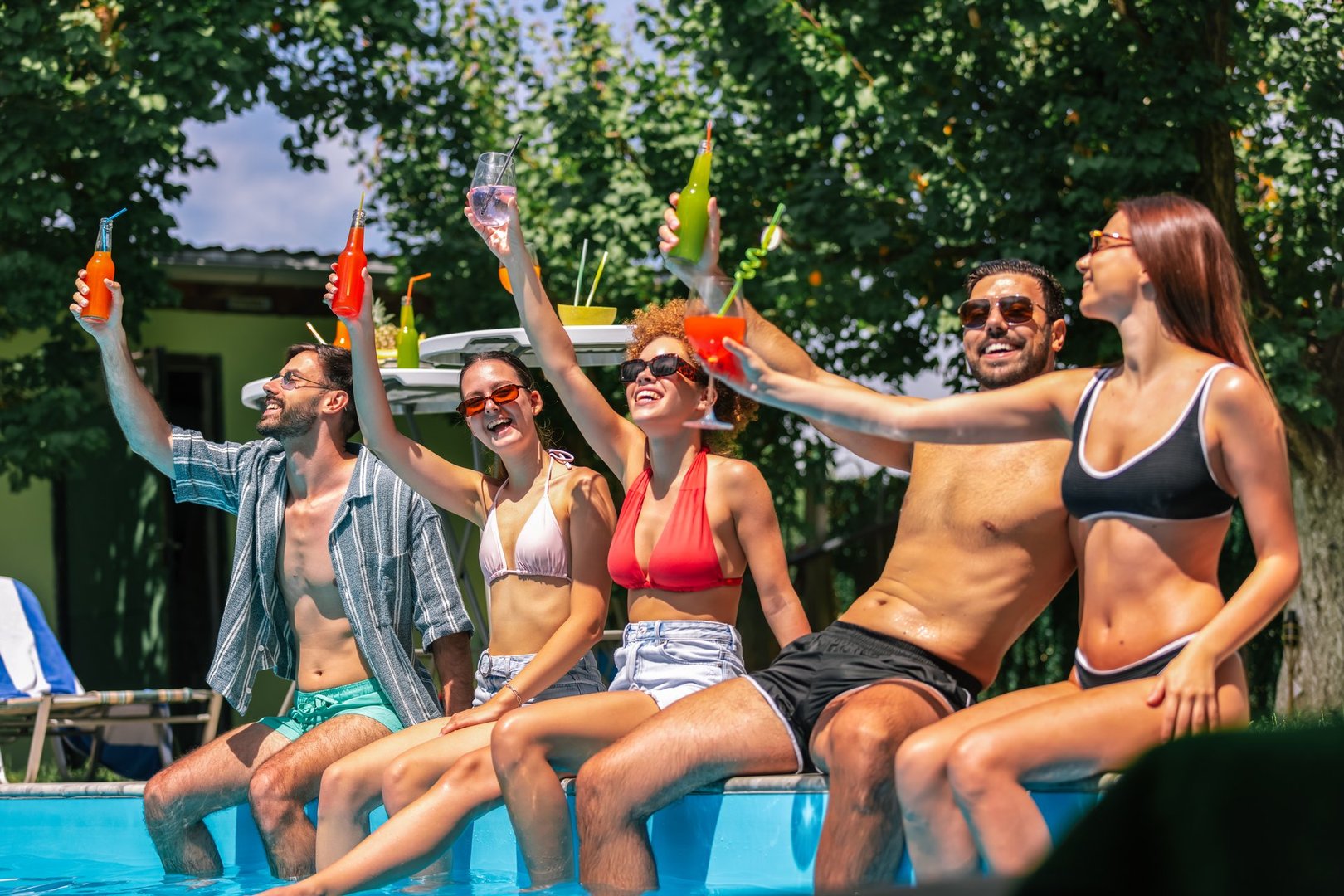 A group of young people in bathing suits sit by the pool with their legs in the water, enjoying colorful cocktails and having a fun, relaxed time together on a sunny day.