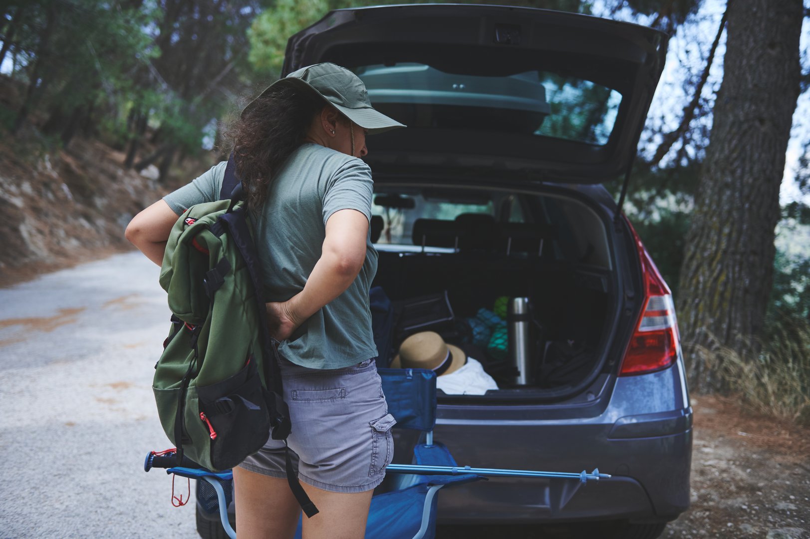 A woman organizes outdoor gear by her open car trunk, suggesting a hiking or camping trip. The wooded background adds a sense of adventure, exploration, and nature.