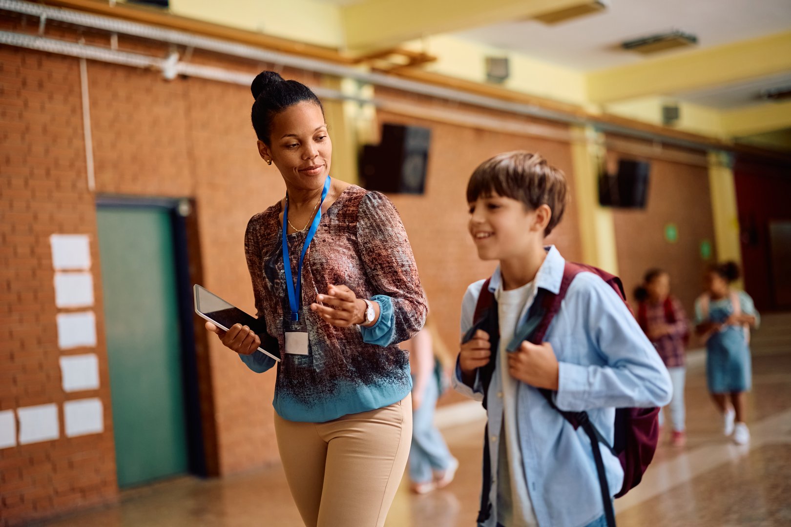 Happy African American teacher and elementary student talking while walking through hallway.