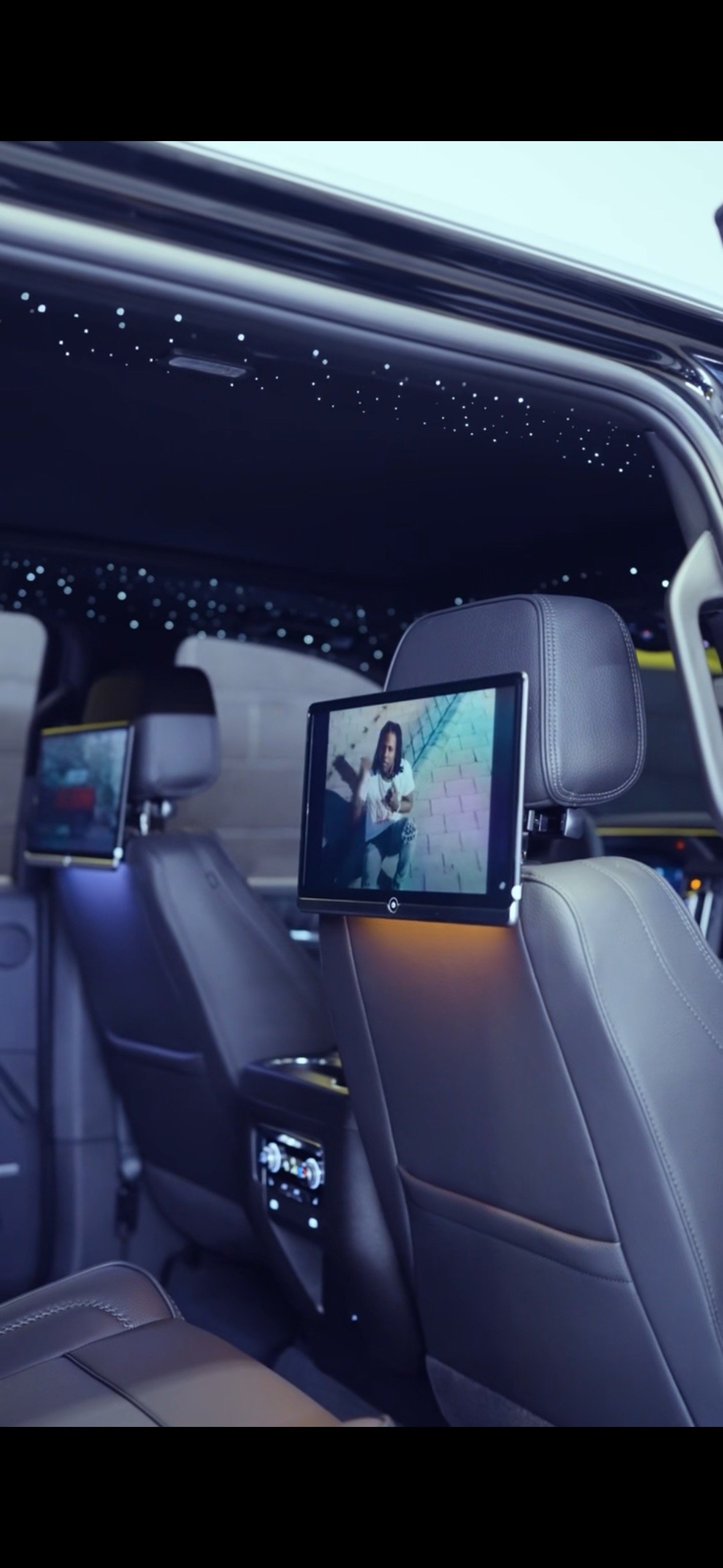 Car interior with two screens on seat backs showing movies, illuminated by star-like ceiling lights.