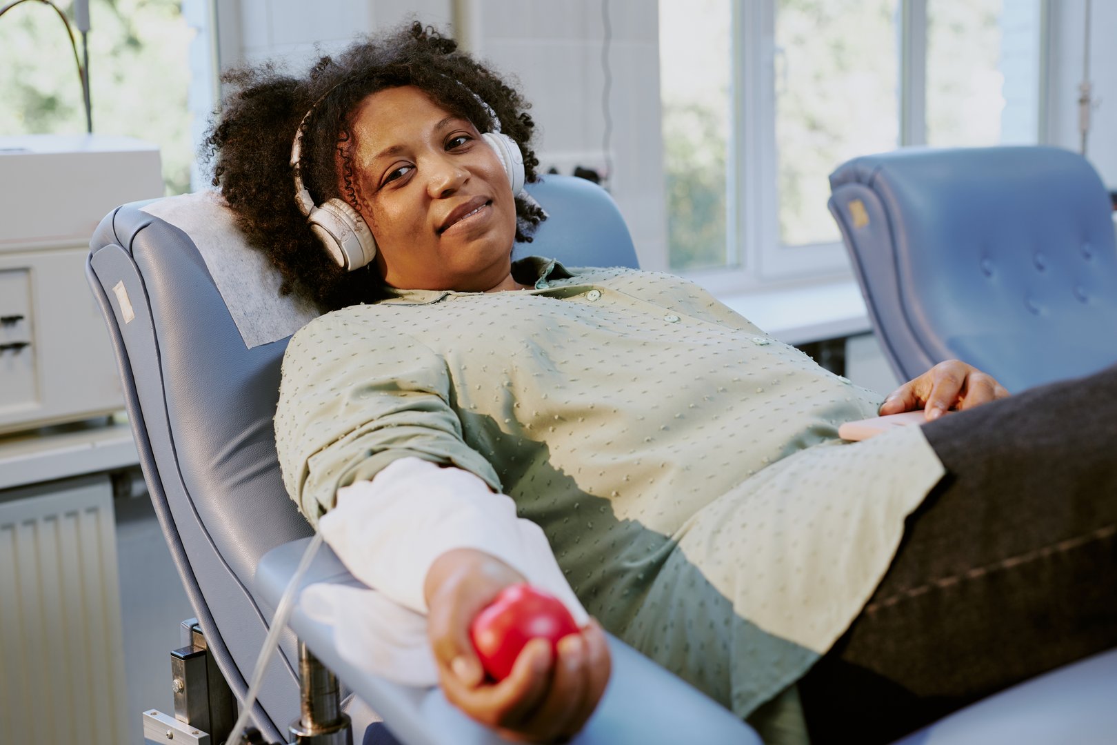 Woman relaxing on donation chair, holding red ball and wearing headphones, during blood donation process in bright medical room