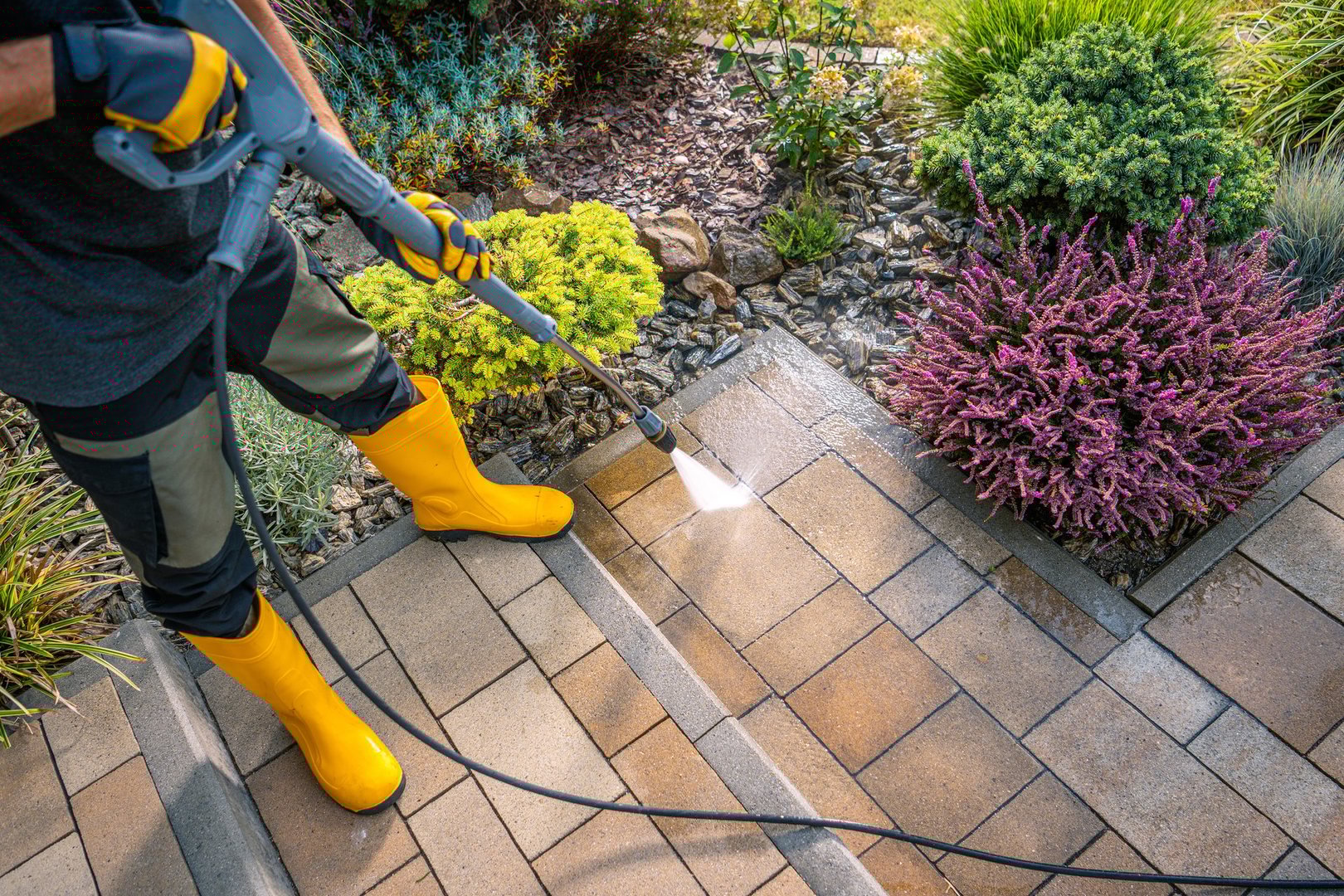A person wearing yellow boots uses a pressure washer to clean paver stones in a well-kept garden filled with vibrant plants and flowers under bright sunlight.