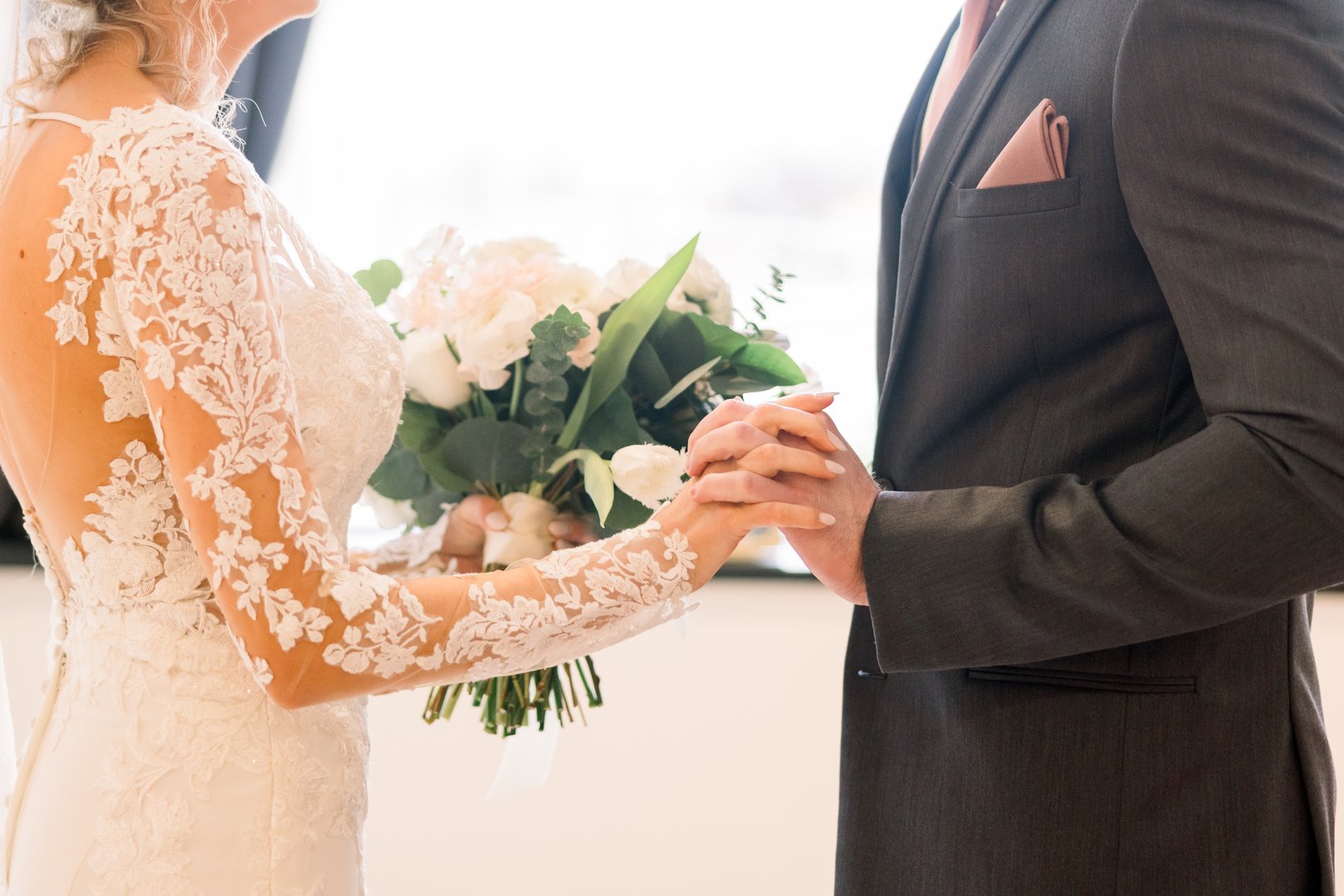 A bride and groom are holding hands in an intimate embrace while she holds her bridal bouquet with her other hand.