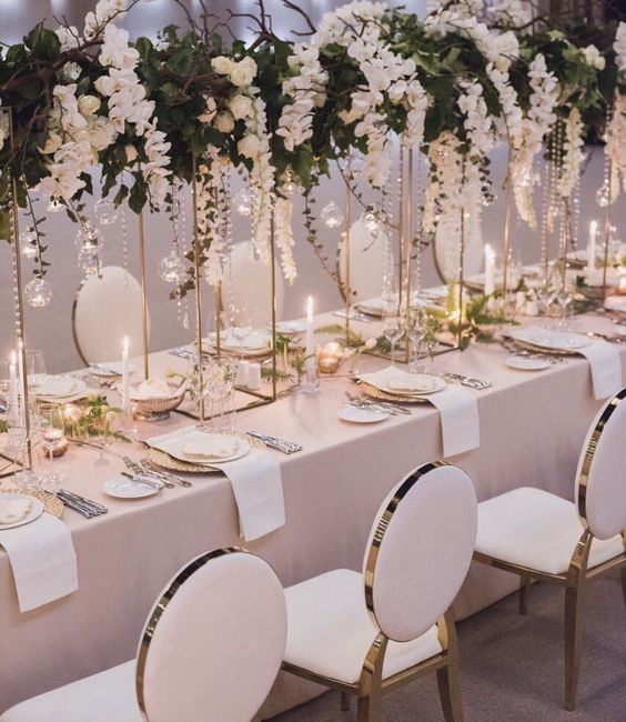 Elegant table setting with white flowers, candles, and pearl décor. Round chairs surround a long, light pink tablecloth.