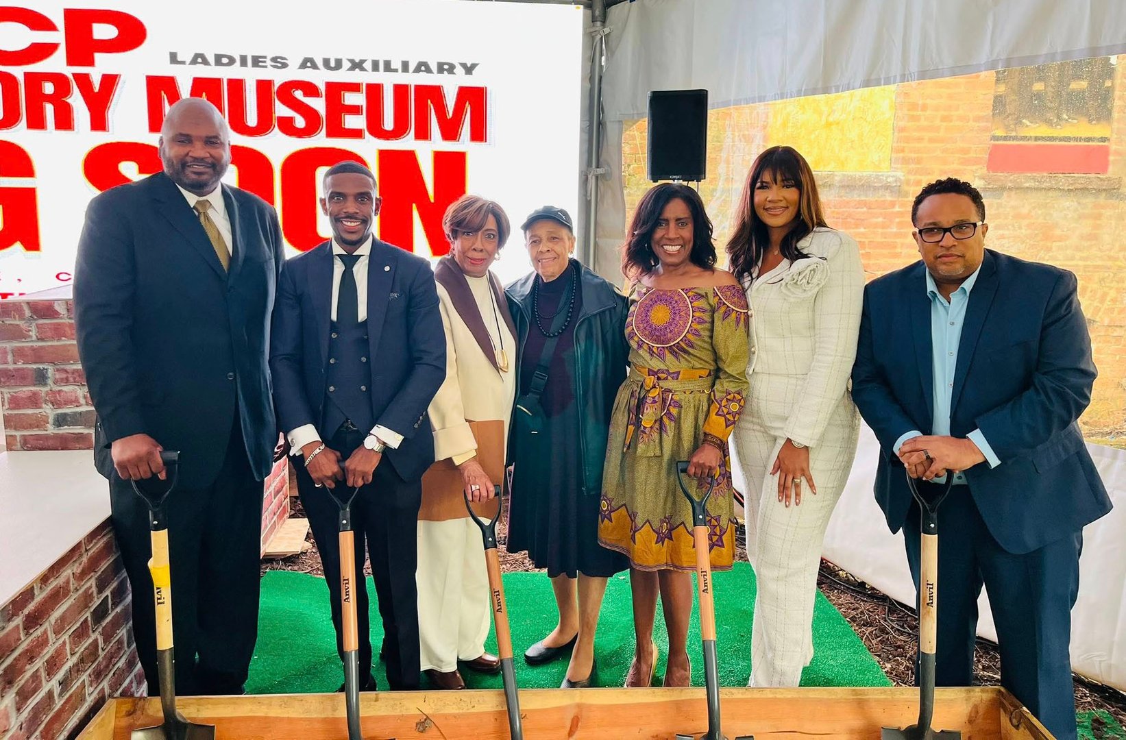 Group of people with shovels at a groundbreaking event for a museum, standing in front of a sign with "COMING SOON."