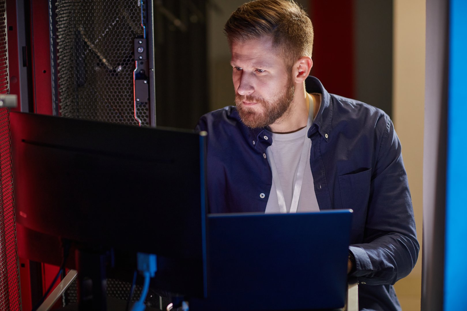 Man with beard observing computer screen in server room setting with technology equipment surrounding him and intense concentration on his face