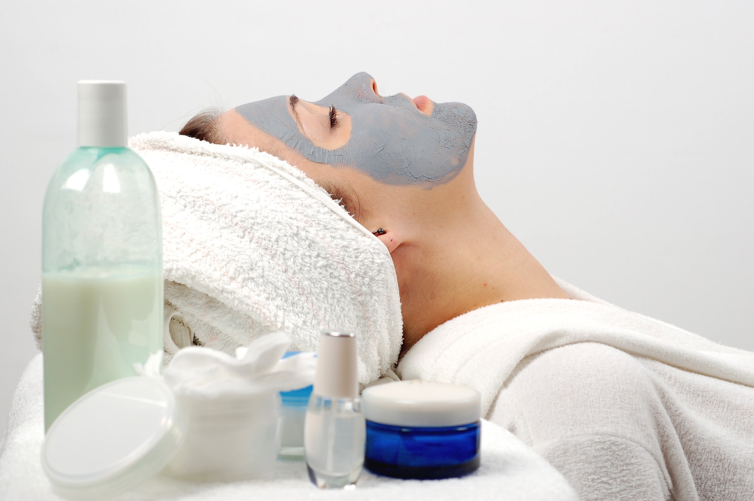 Person with a grey face mask and towel on head, surrounded by skincare products, relaxing in a spa.