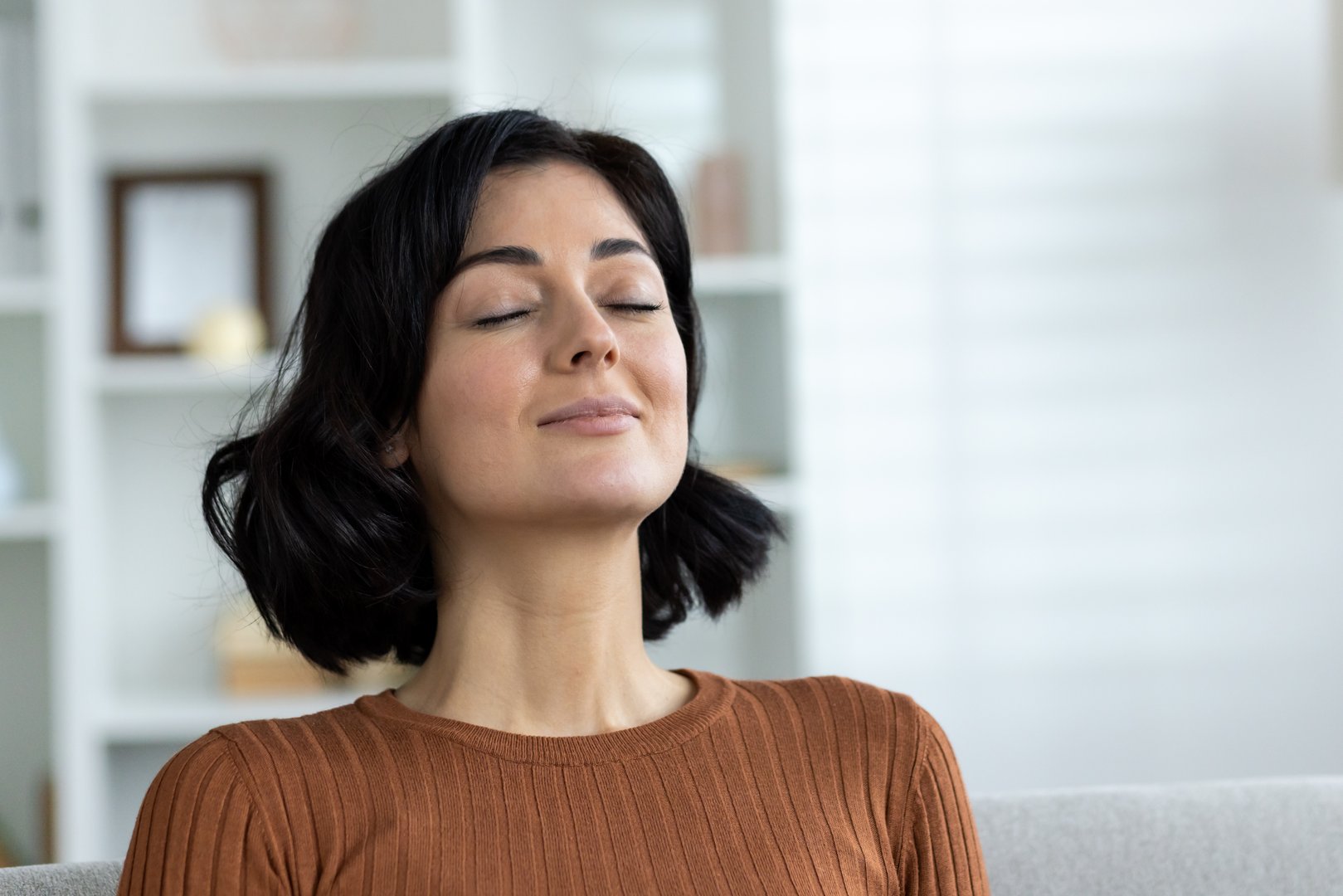 Woman with short dark hair, eyes closed, meditating and practicing mindfulness at home, relaxing on a couch in a cozy, bright room.