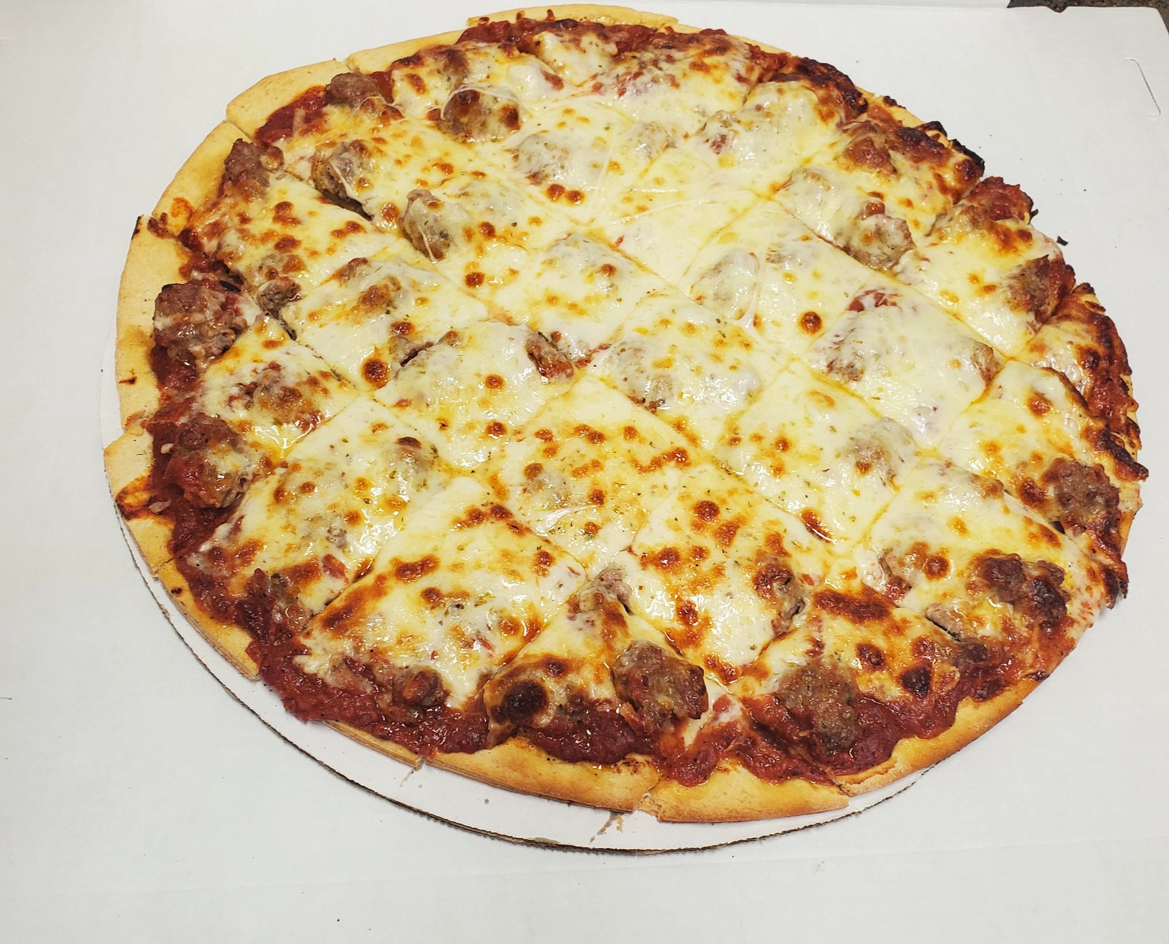 A sliced pizza with melted cheese and sausage on a thin crust, placed on a round cardboard base.