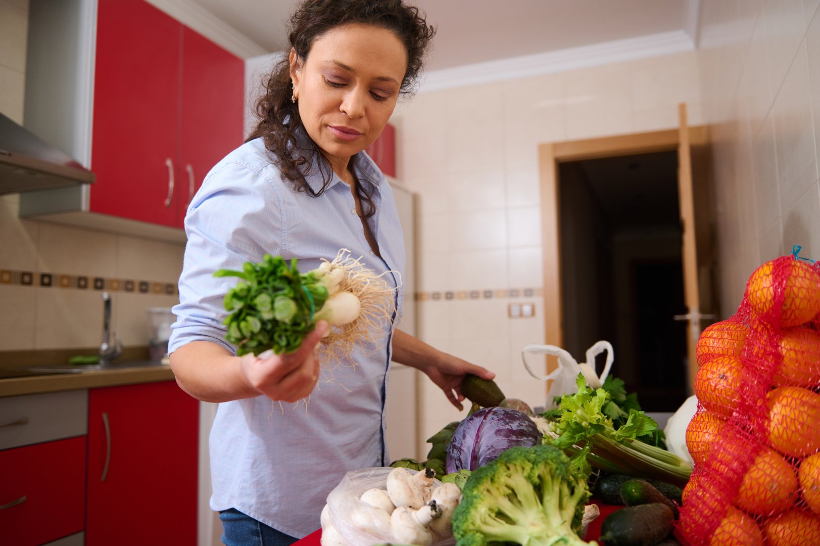 A woman in a modern kitchen sorts through fresh vegetables, surrounded by greens, oranges, and mushrooms. The setting suggests a focus on healthy, home-cooked meals with natural ingredients - High Performance Nutrition at SugarFree.Space
