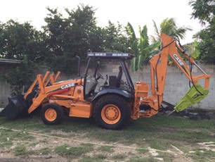 An orange CASE backhoe loader parked on grass with trees and a wall in the background.