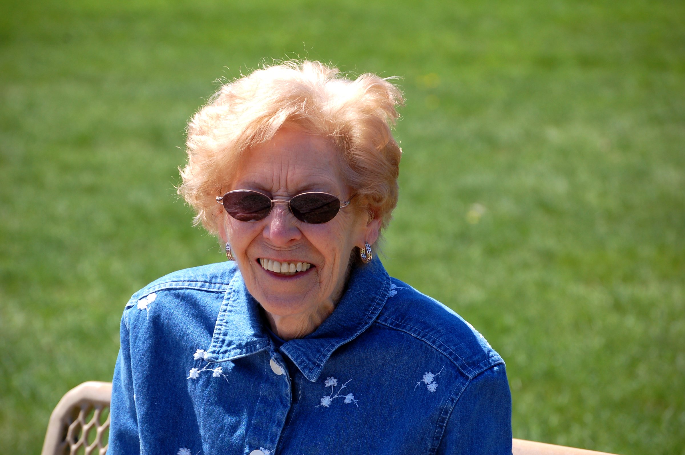 Elderly woman with sunglasses smiles, wearing a denim jacket with floral embroidery, seated outdoors on a sunny day.