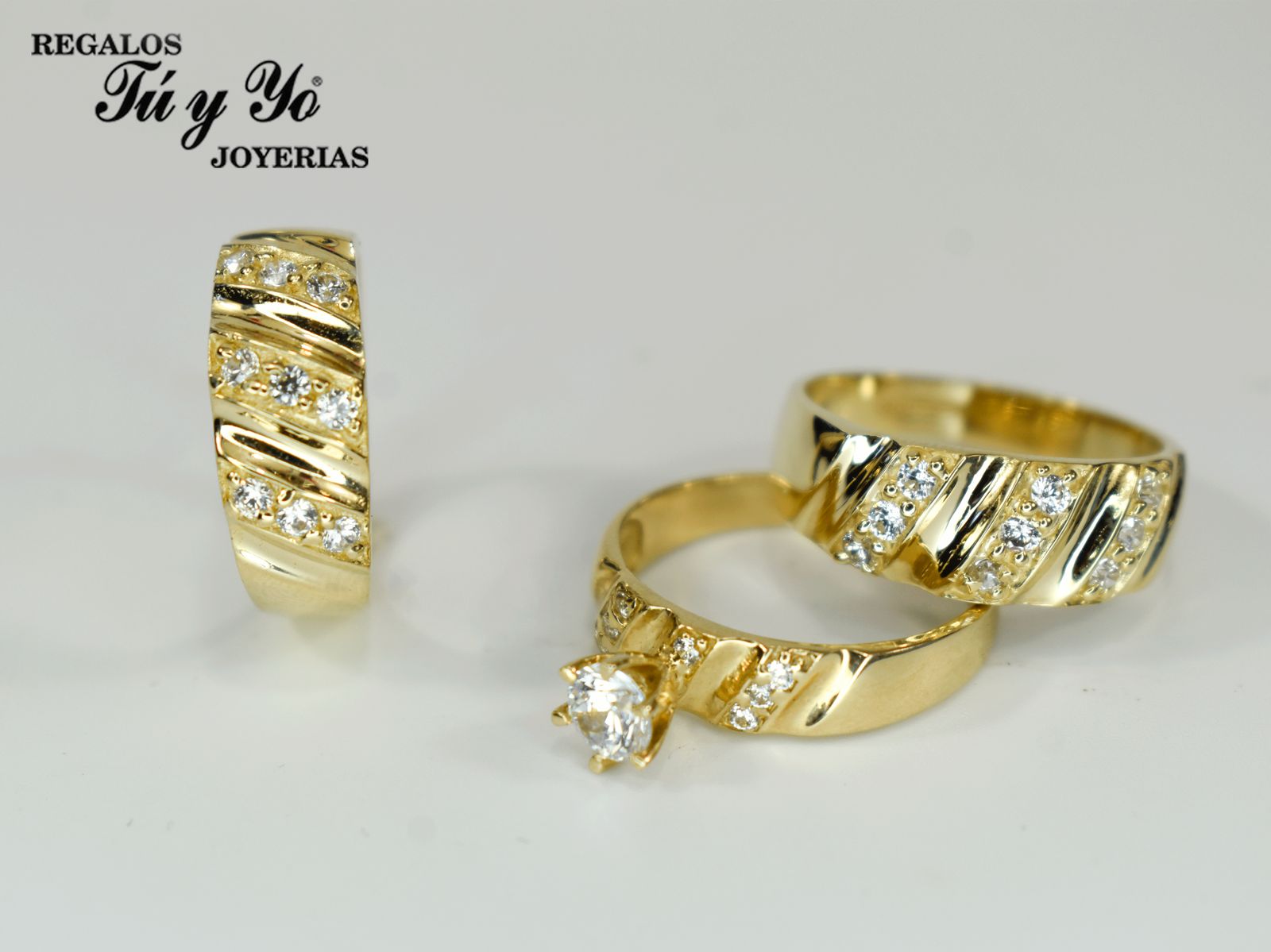 Three gold rings with diamond accents displayed on a white surface, featuring branding for "Regalos Tú y Yo Joyerías."