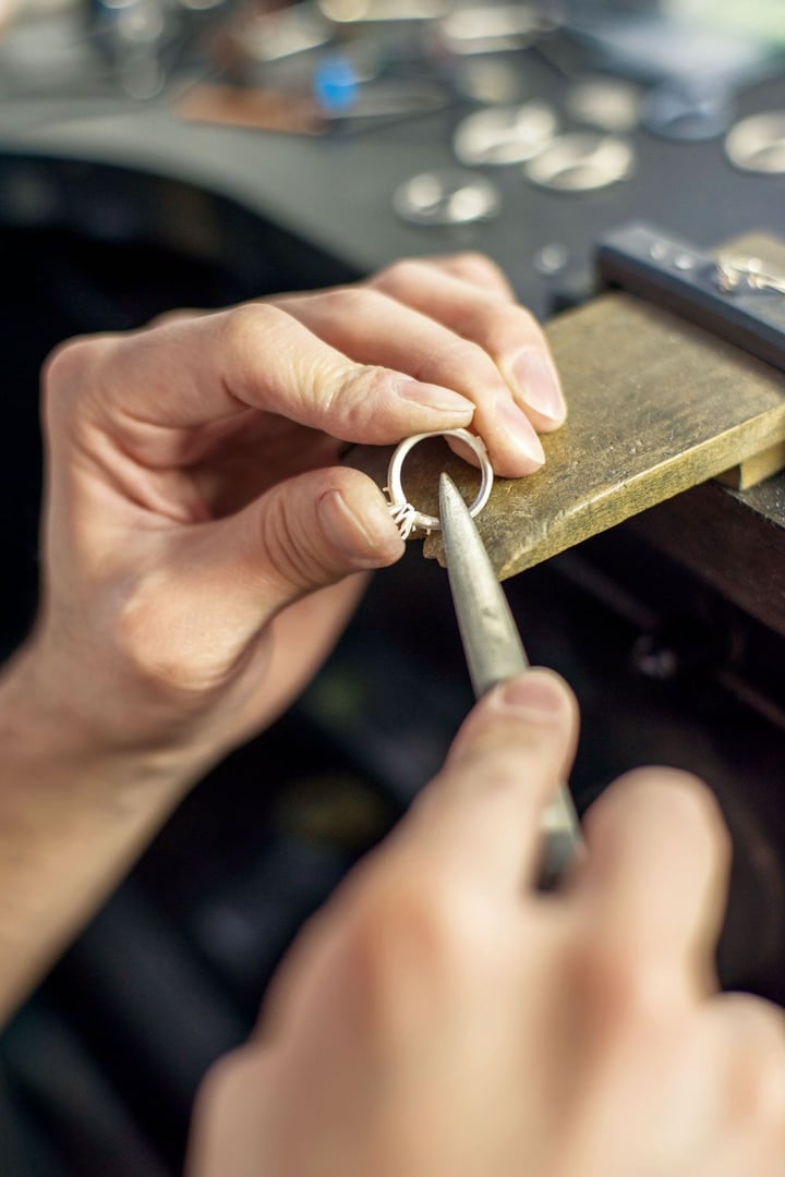 Person using a tool to craft or repair a silver ring at a jewelry workbench.