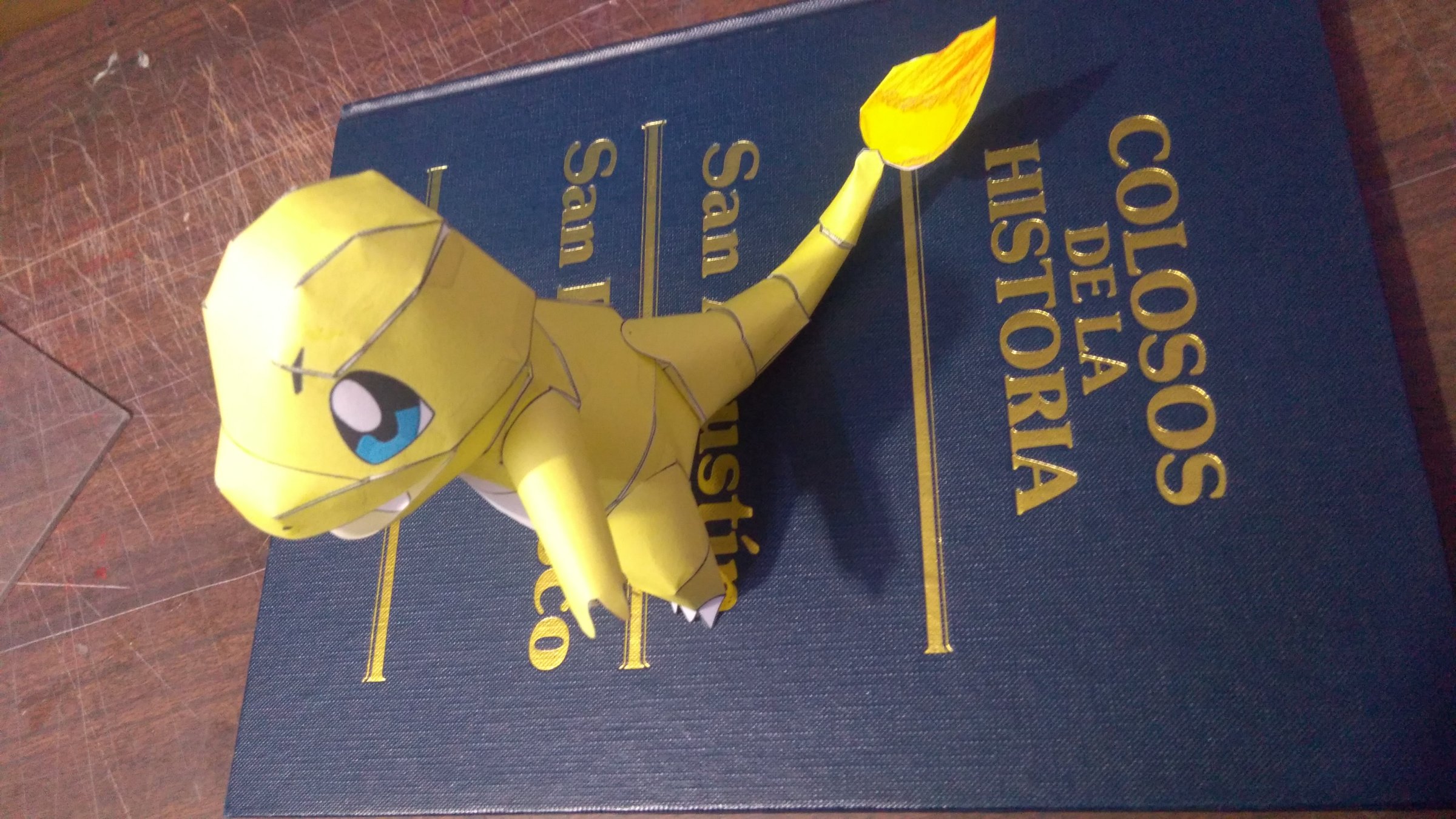 A yellow paper model of a dinosaur-like creature resting on a blue and gold book titled "Colosos de la Historia."