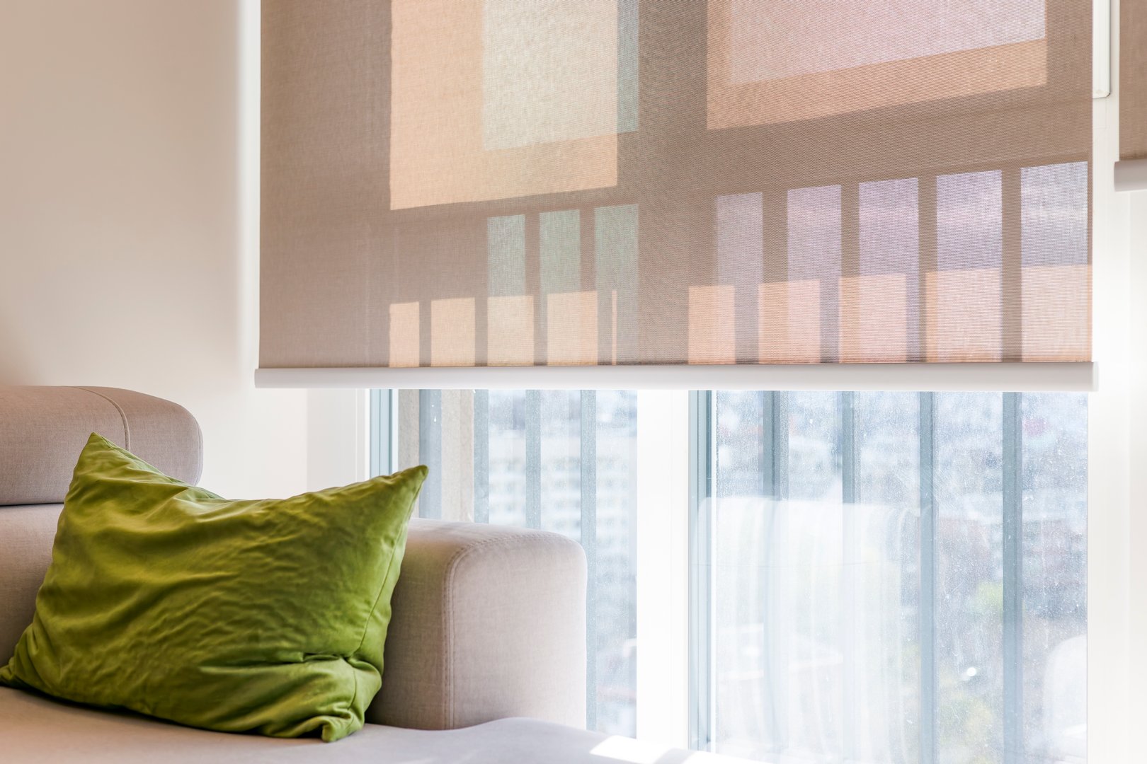 Beige roller blinds, close-up view, covers a large window, softly filtering sunlight and casting geometric shadows. Sunscreen fabric of roller shades creates a cozy feel in the bright interior space.