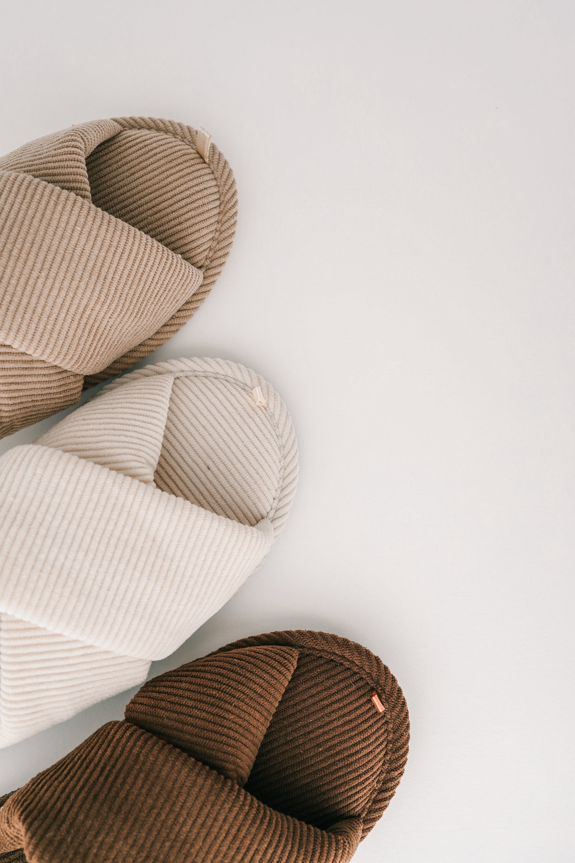 Three pairs of ribbed fabric slippers in neutral tones-beige, white, and brown-are arranged on a clean white background. The minimalist and cozy design emphasizes comfort and modern home aesthetics.