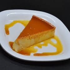 A slice of caramel flan on a white plate, drizzled with caramel sauce, set against a dark background.