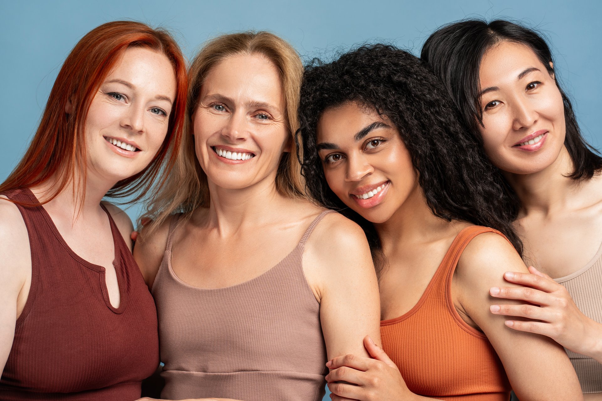 Four diverse, multinational women, fashion modes embracing, looking at camera, closeup posing in studio isolated on blue background. Advertisement concept