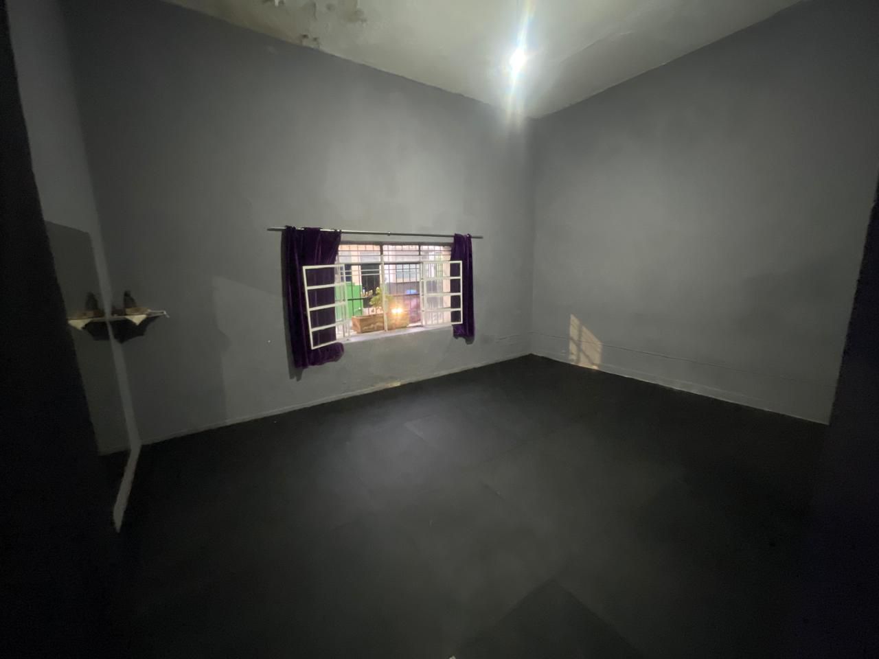 A dimly lit empty room with gray walls and black floor tiles, featuring a window with bars and curtains, and a small wall shelf.