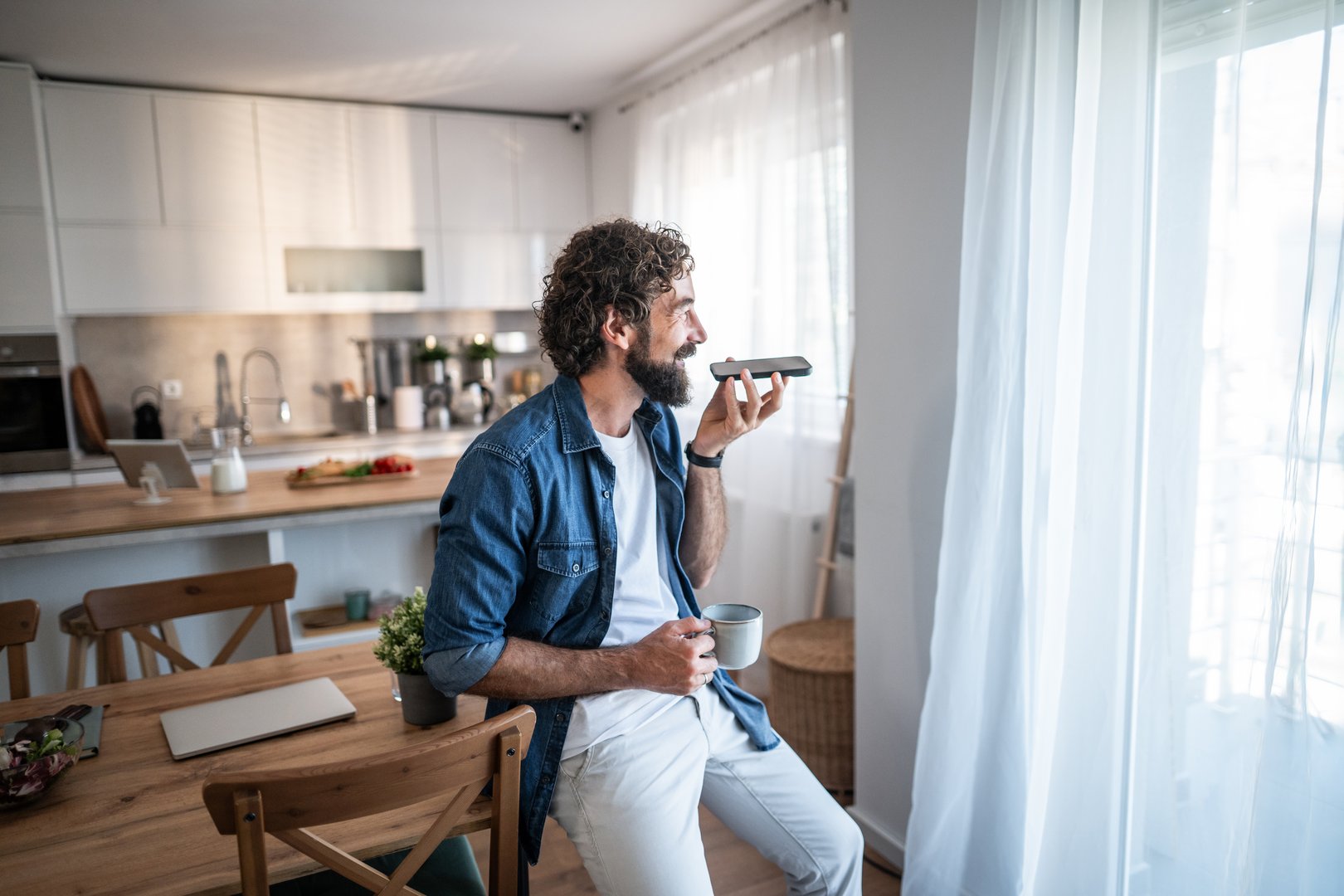 Man holding a coffee mug, leaning on a table in his kitchen and speaking into a smartphone on speakerphone, utilizing voice command technology for hands-free communication
