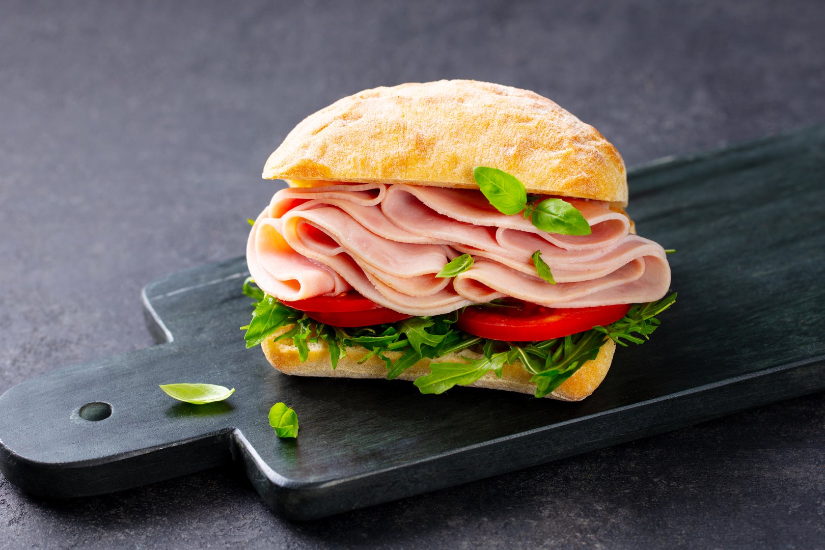 Ciabatta sandwich with ham, fresh tomatoes and green salad on a marble board. Dark background. Close up.