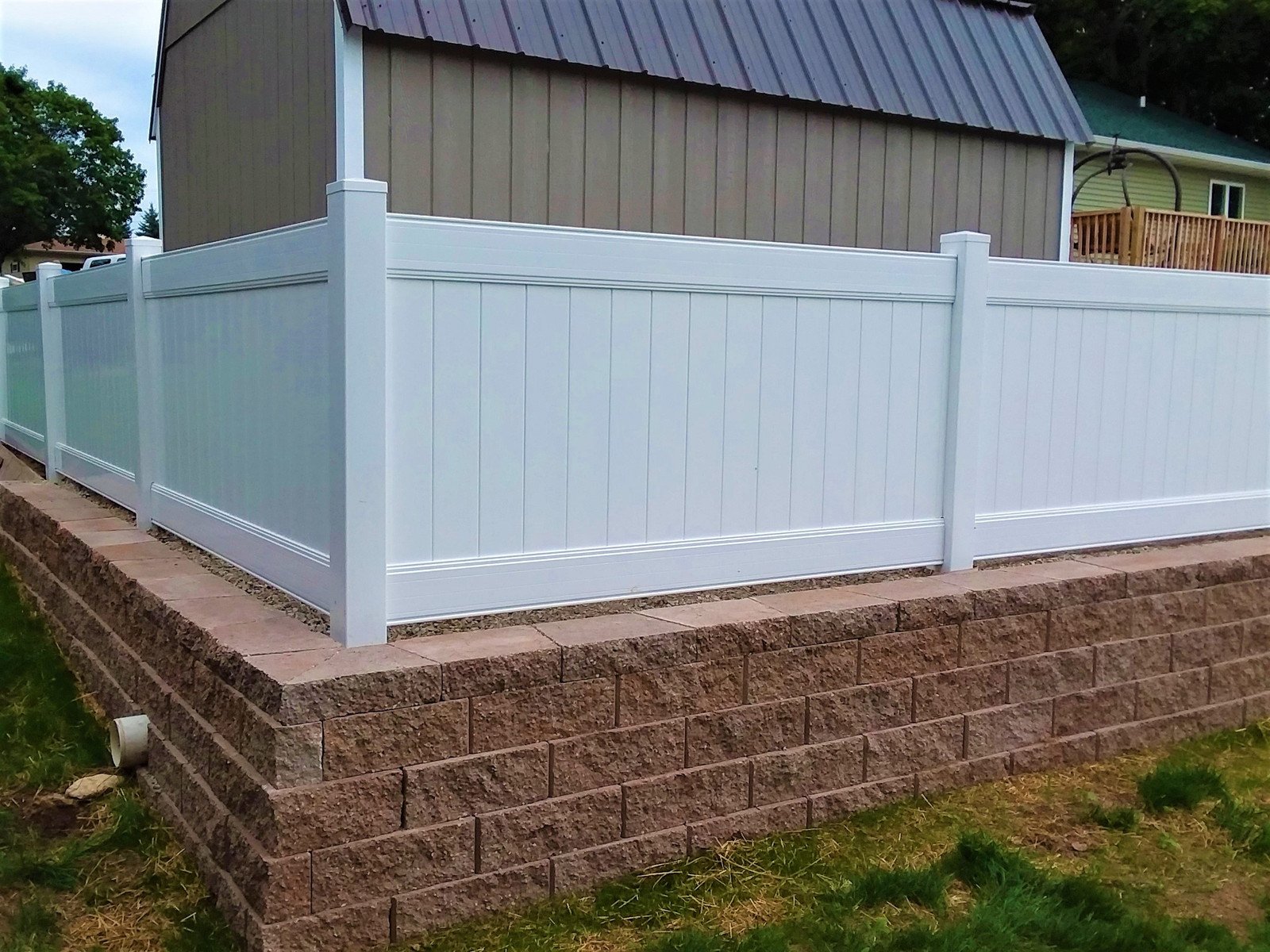 A white vinyl privacy fence on top of a stone retaining wall in a residential backyard.