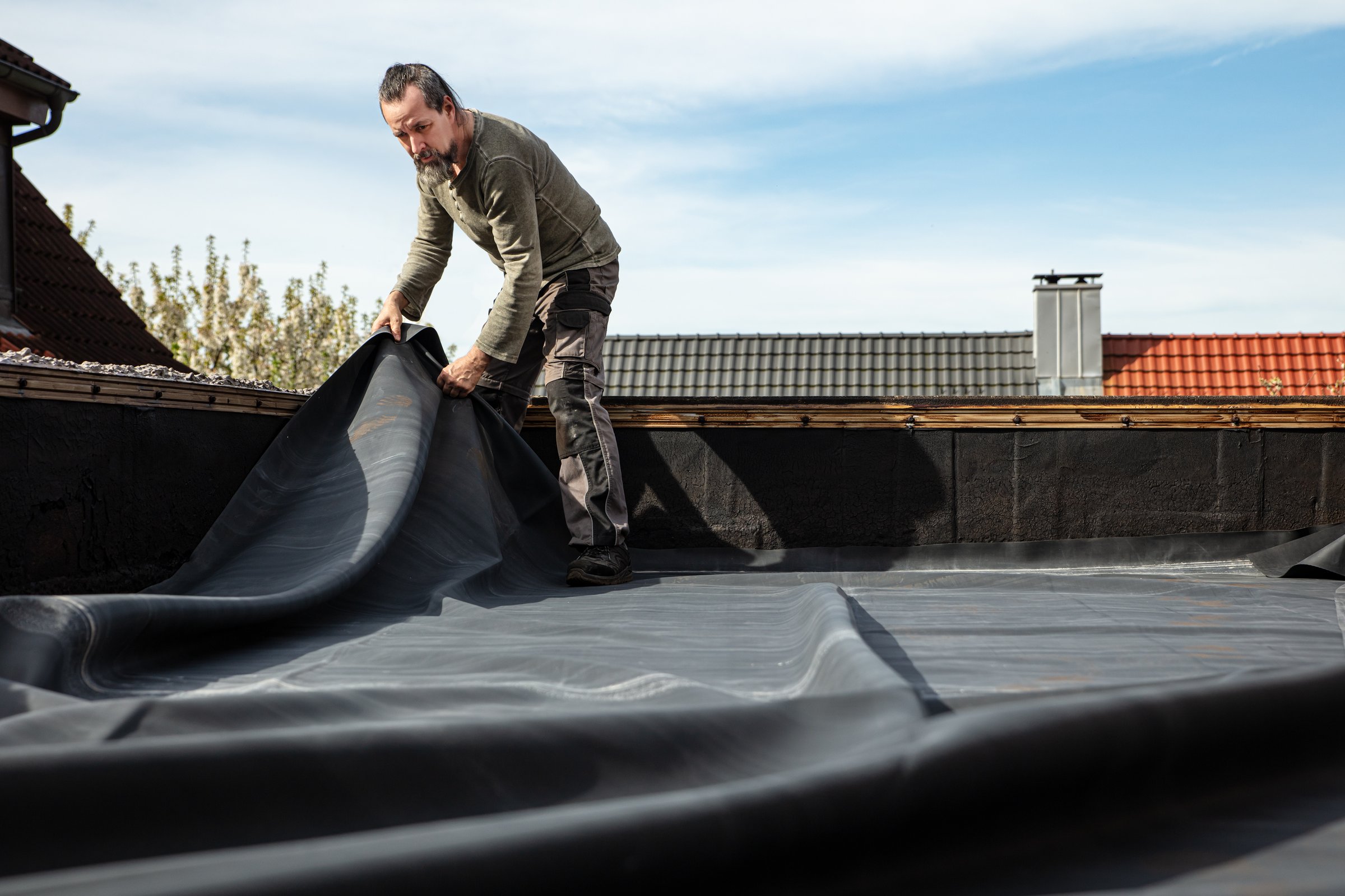 A craftsman rolls out a thick rubber tarpaulin on a flat roof to re-seal and repair the broken bitumen coating