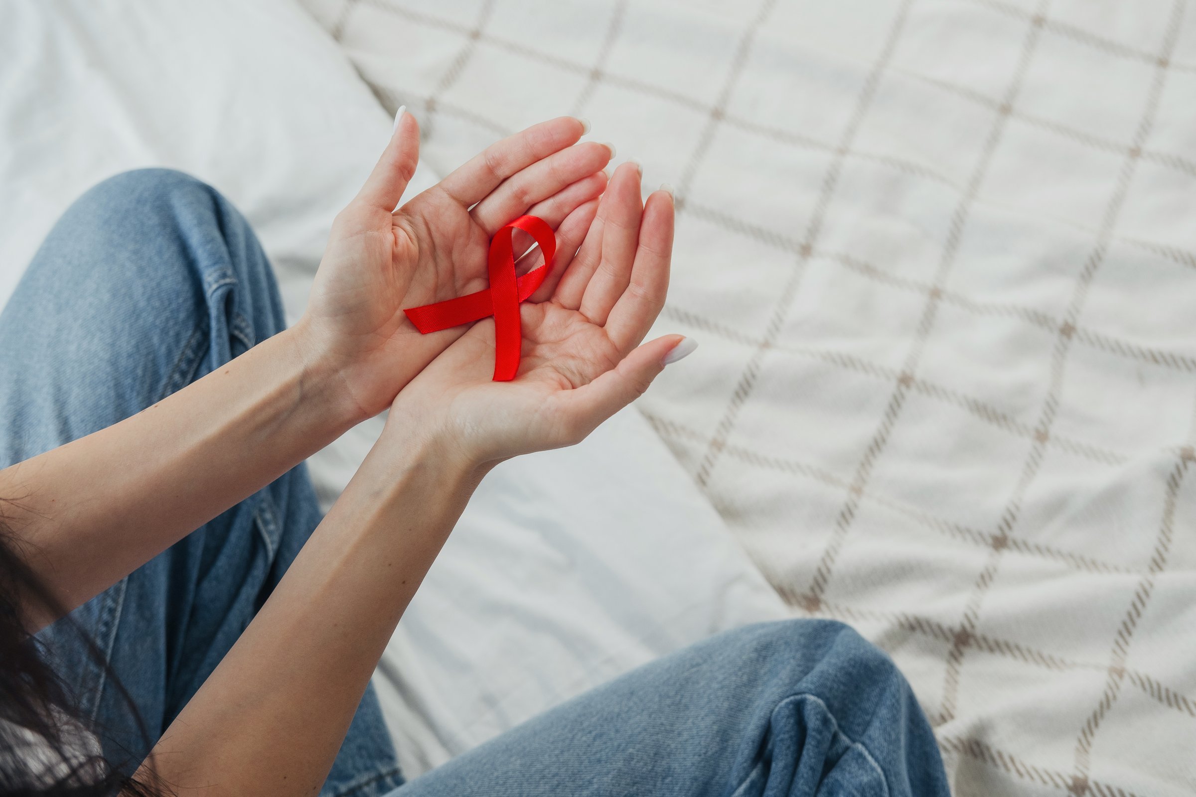 A person gently holds a red ribbon in their palms, sitting on a bed with a checkered design.