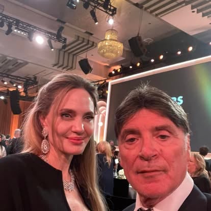 Two people pose for a selfie at an indoor event with a chandelier and a screen in the background.