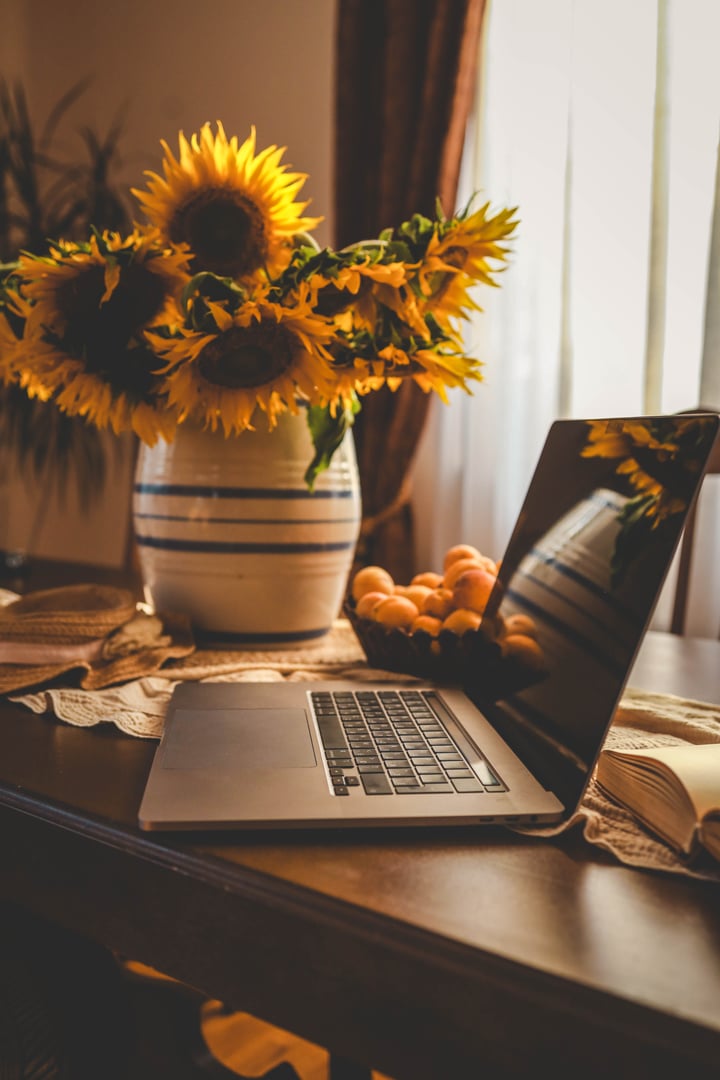A serene scene featuring a table with a vase of sunflowers, a laptop, a bowl of apricots, and an open book. Soft daylight filters through the curtains, casting a warm and cozy ambiance.