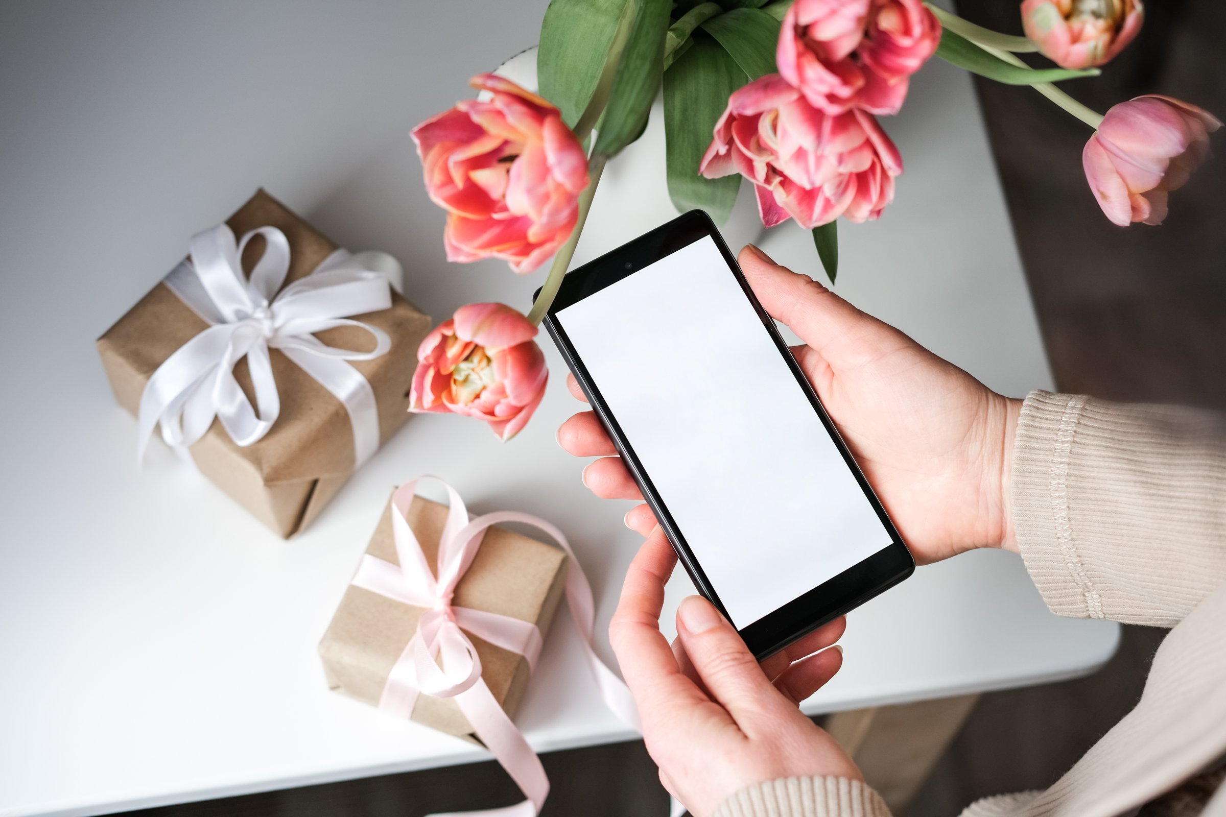 Woman holding smartphone with blank white screen over white table with two wrapped gifts and bouquet of pink tulips. Online gift shopping concept