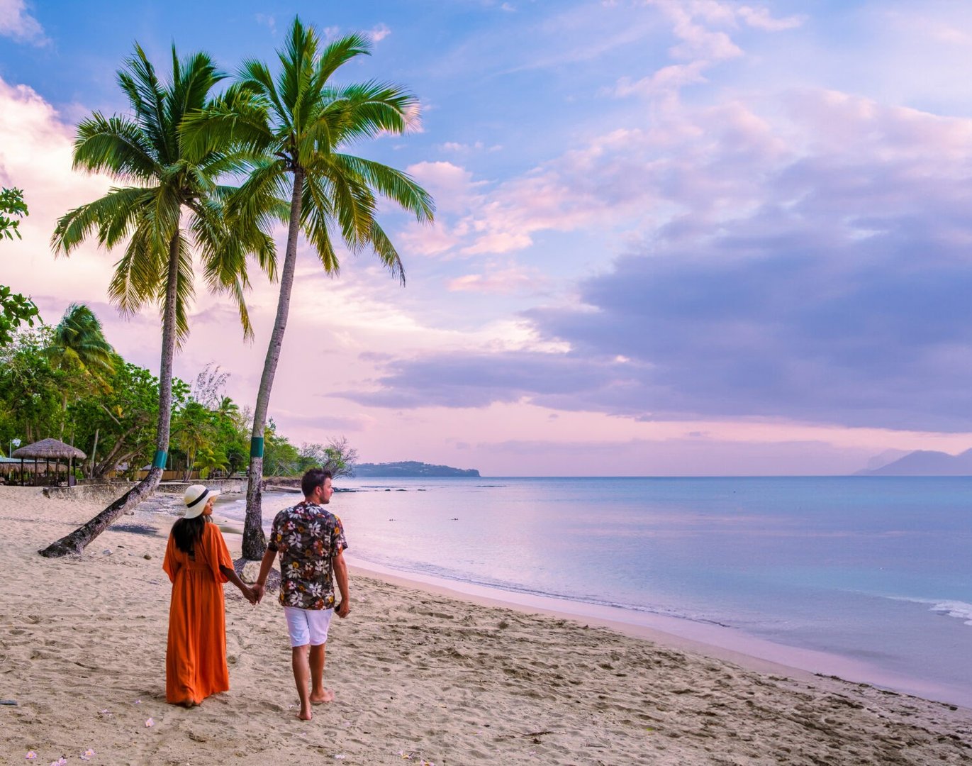 A couple holding hands walks along a tropical beach at sunset, with palm trees and calm ocean waves in the background.