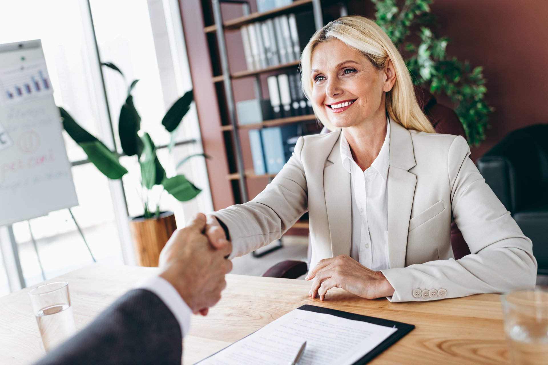 A female business professional in formal attire shakes hands with a colleague across a desk, symbolizing agreement, partnership, and teamwork in a professional corporate setting.