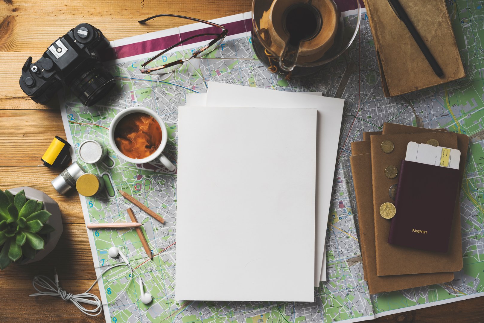 Travel concept flat lay table top shot on wooden background. Map, coffee, passport, glasses, earphones, succulent, pencils, notebook, camera and film rolls on the table with white blank cover magazine stack.