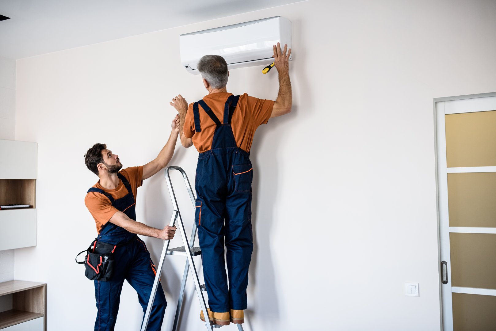 Two workers in uniform, air conditioning masters using ladder while installing a new air conditioner in the apartment. Construction, maintenance and repair concept. Horizontal shot