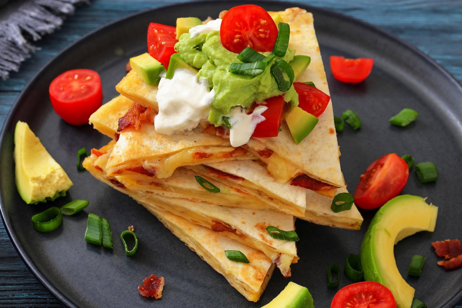 stack of cut up quesadilla with cheese and fried bacon topped with sour cream, guacamole, avocado and tomatoes on a plate on wood table with ingredients, not AI generated, close-up