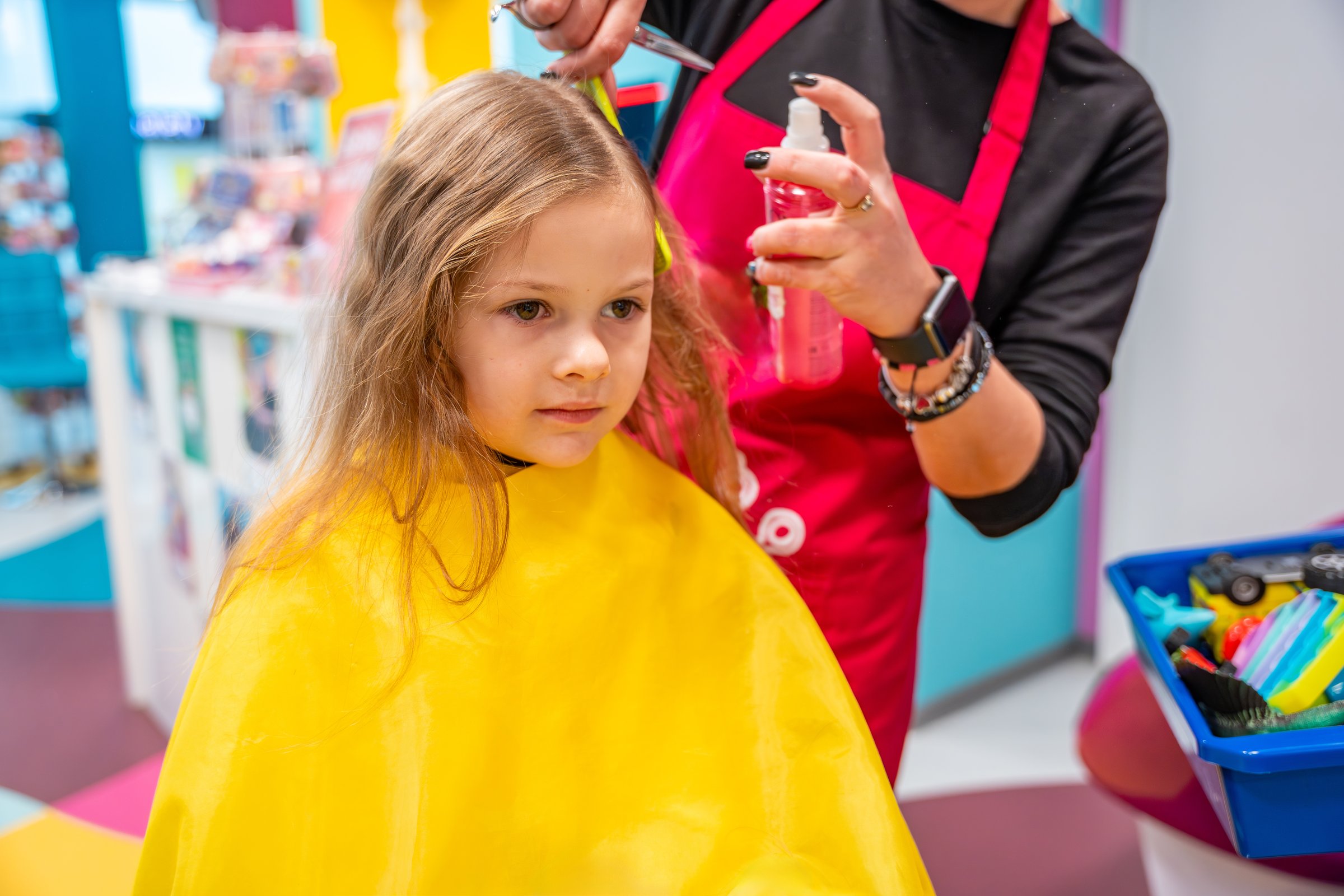 Hairdresser making a hair style to cute little girl in real child salon with colorful interior and toys. High quality photo