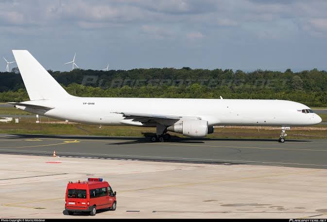 White aircraft on a runway with a red vehicle nearby, set against a grassy background with wind turbines in the distance.