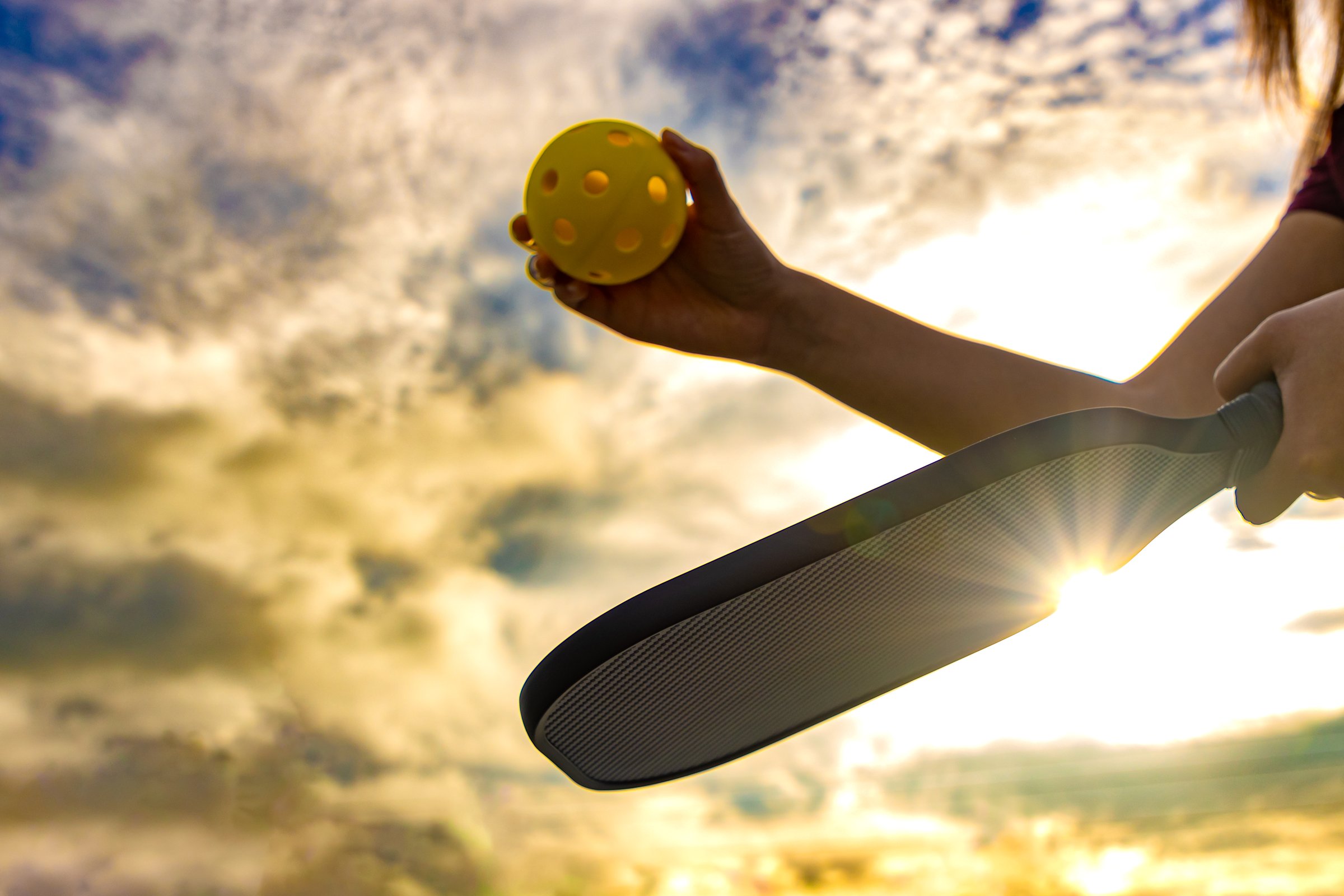 A woman prepares to serve during a game of Pickleball by holding the ball above her paddle as the sun sets or rises in the background.