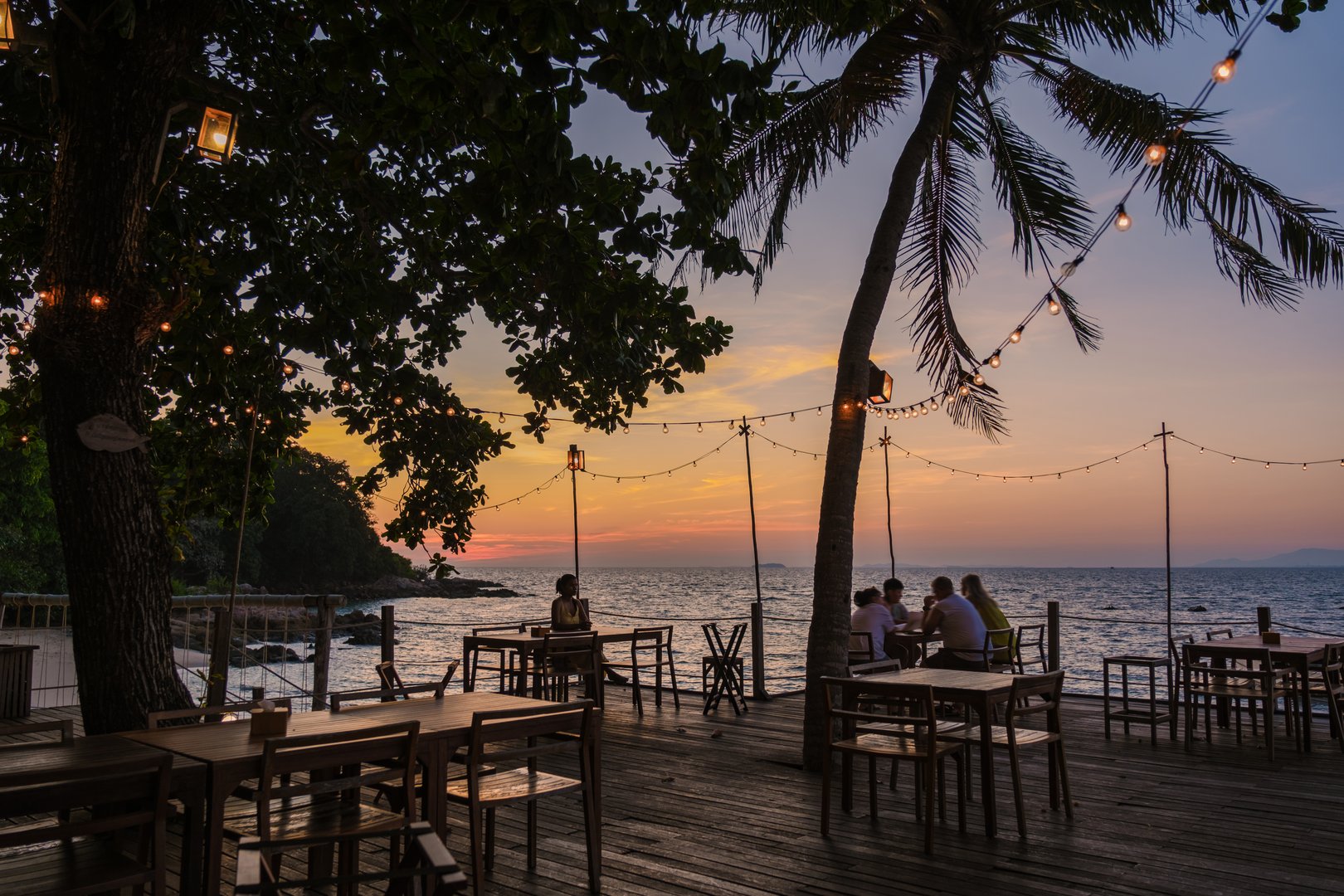 Guests enjoy a tranquil evening at a beachside restaurant on Koh Munnork Island, surrounded by lush trees and twinkling lights as the sun sets over the calm waves of the Gulf of Thailand.