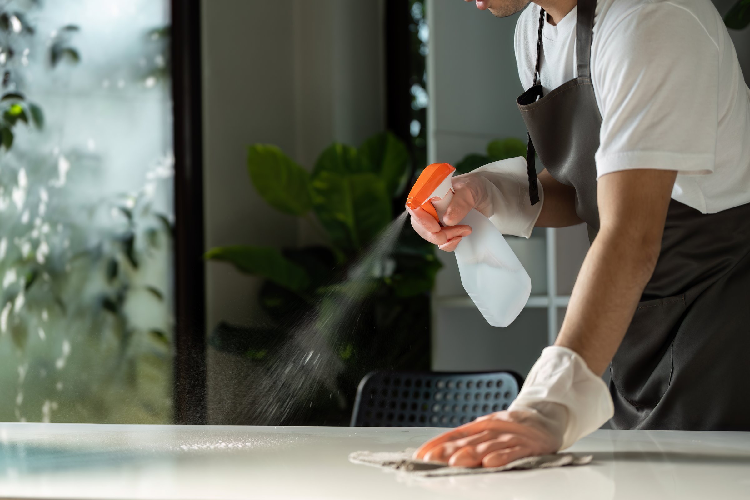 A person wearing gloves cleans a kitchen countertop with a spray bottle and cloth, emphasizing cleanliness and hygiene.
