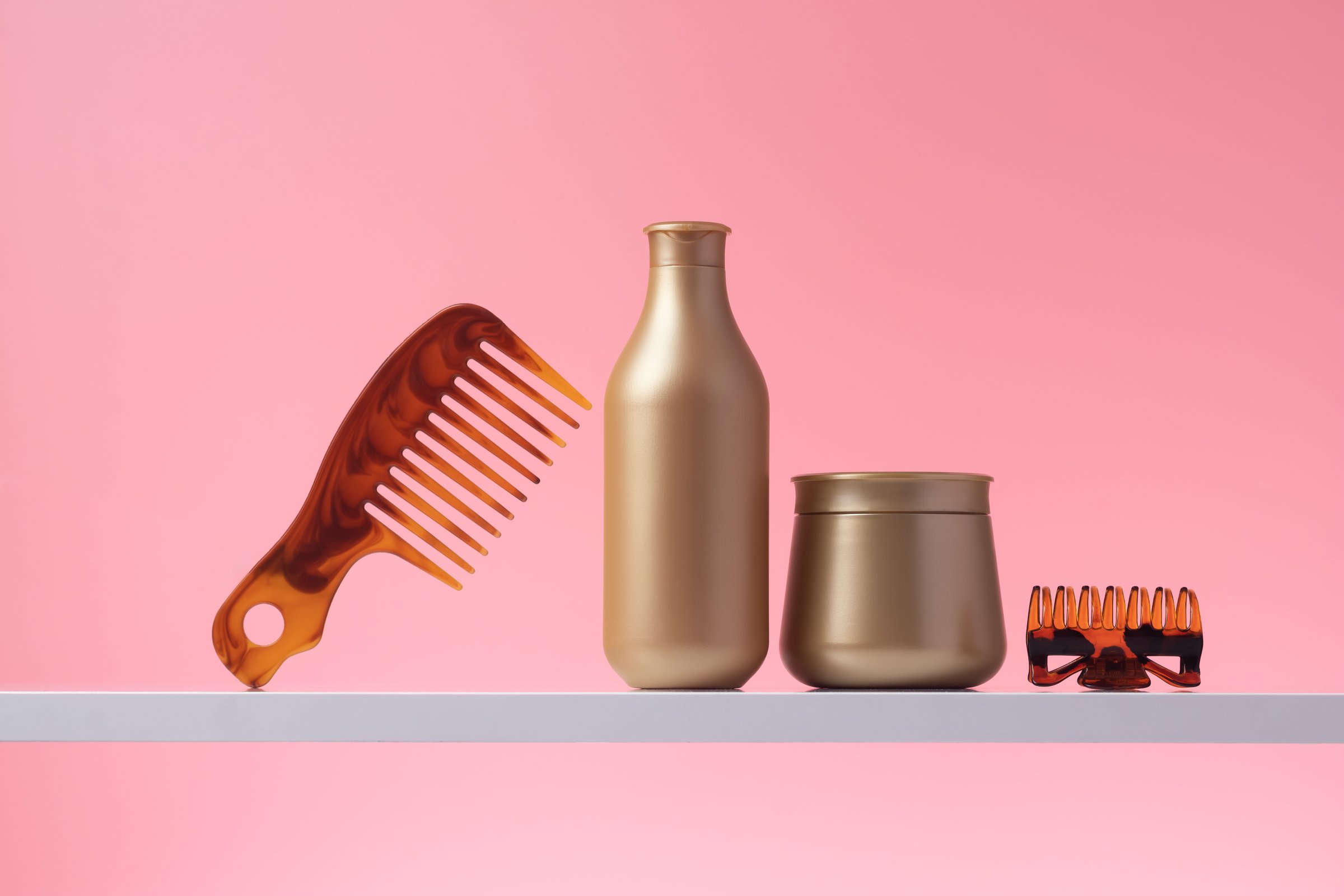 Shampoo and hair conditioner, various hair beauty care products on shelf in home bath, pink background. Comb and gold plastic bottles in bathroom, accessories and toiletries cosmetics. Space for text