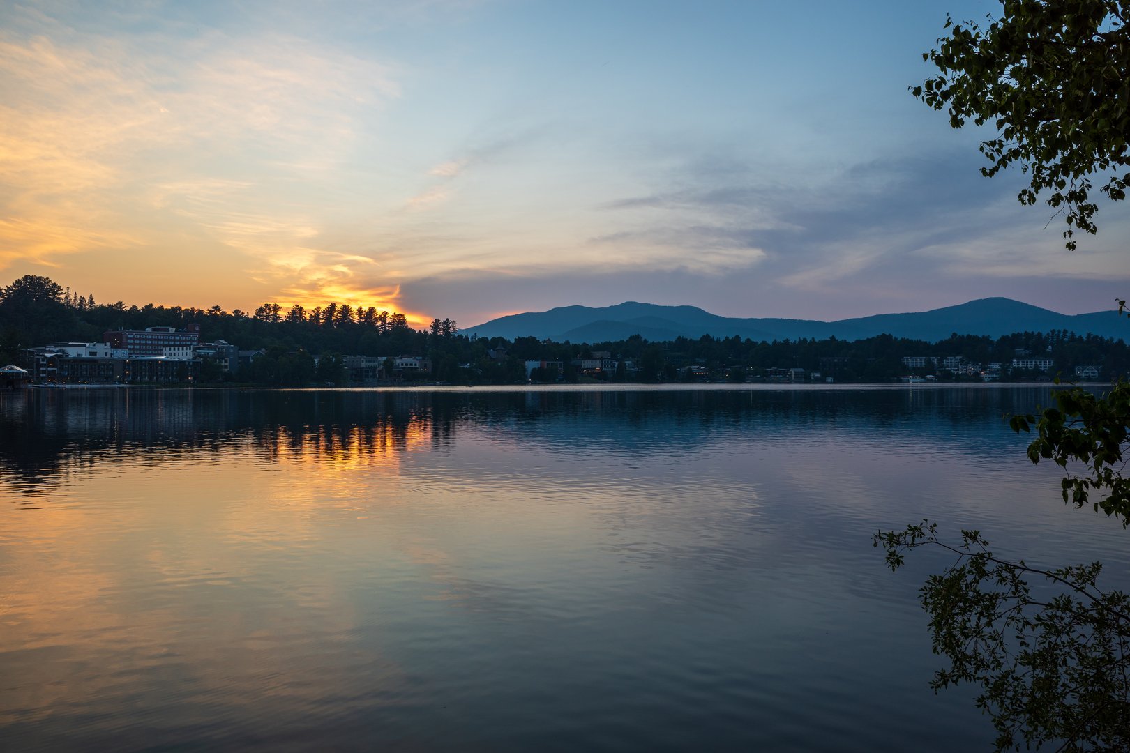 A breathtaking sunset casts golden hues over Lake Placid, reflecting the tranquil beauty of the Adirondack Mountains across Mirror Lake, capturing the serene essence of Upstate New York.
