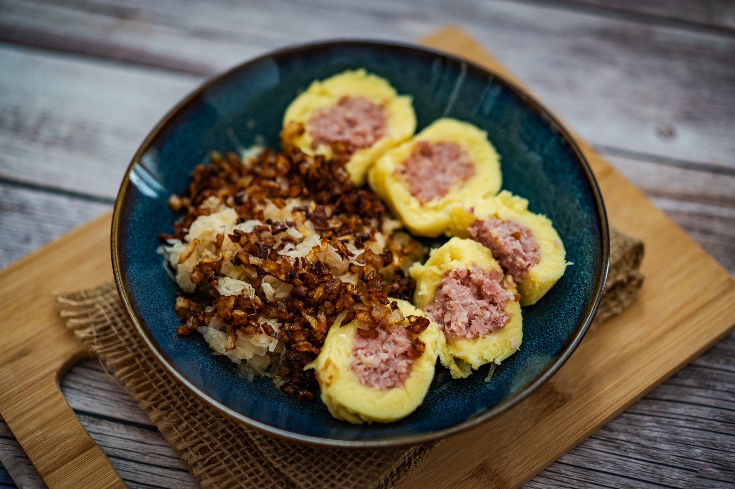 The scene features a traditional Czech dish consisting of potato dumplings filled with smoked meat. Accompanying the dumplings are fermented sauerkraut and crispy fried onions. This classic meal showcases the rich flavors of Czech cuisine.