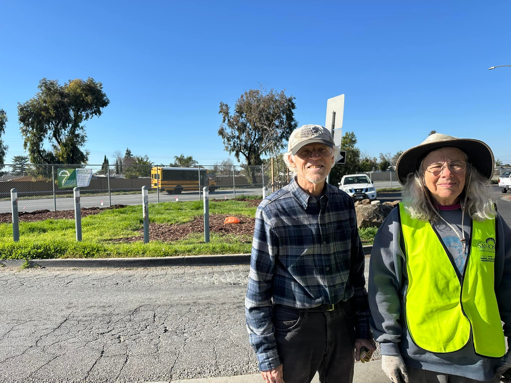 Two older adults in casual clothes and hats stand outdoors on a sunny day with trees and a road in the background.