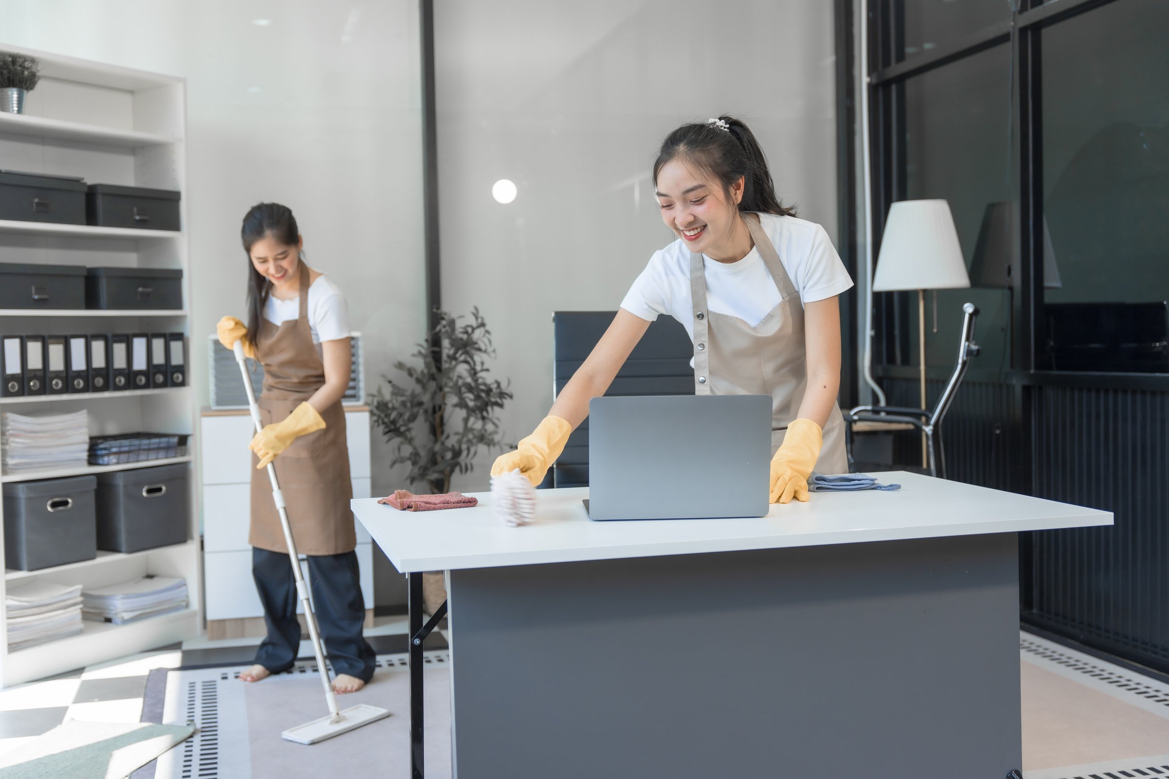 Two Asian housekeepers wear overalls and work together efficiently to clean the living room, study, office.They wipe the windows, mop the floors, inspect the shop to make sure the work is spotless