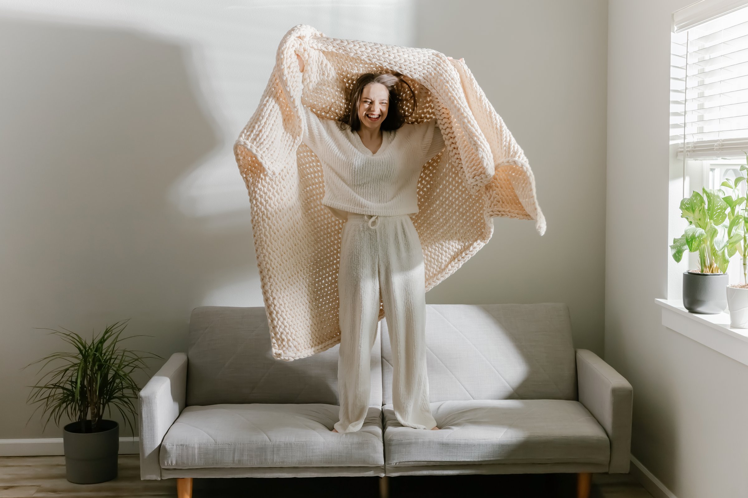 Young woman smiling and holding a chunky knit blanket above her while standing on a sofa in soft loungewear. A fun and carefree cozy moment at home.